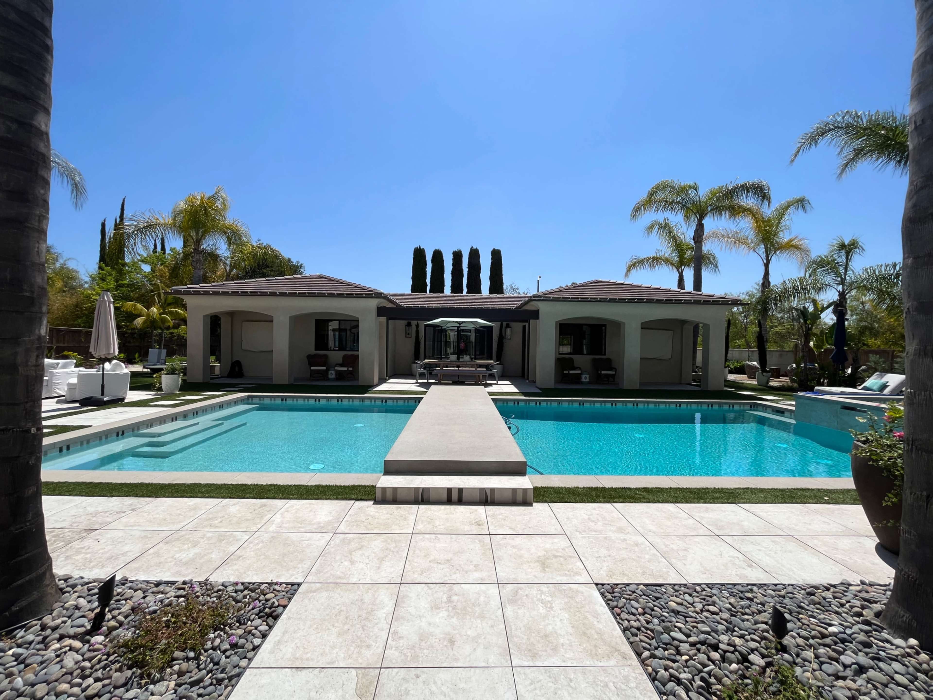 A rectangular swimming pool is bordered by a stone patio and leads to a single-story house with multiple palm trees in the background.