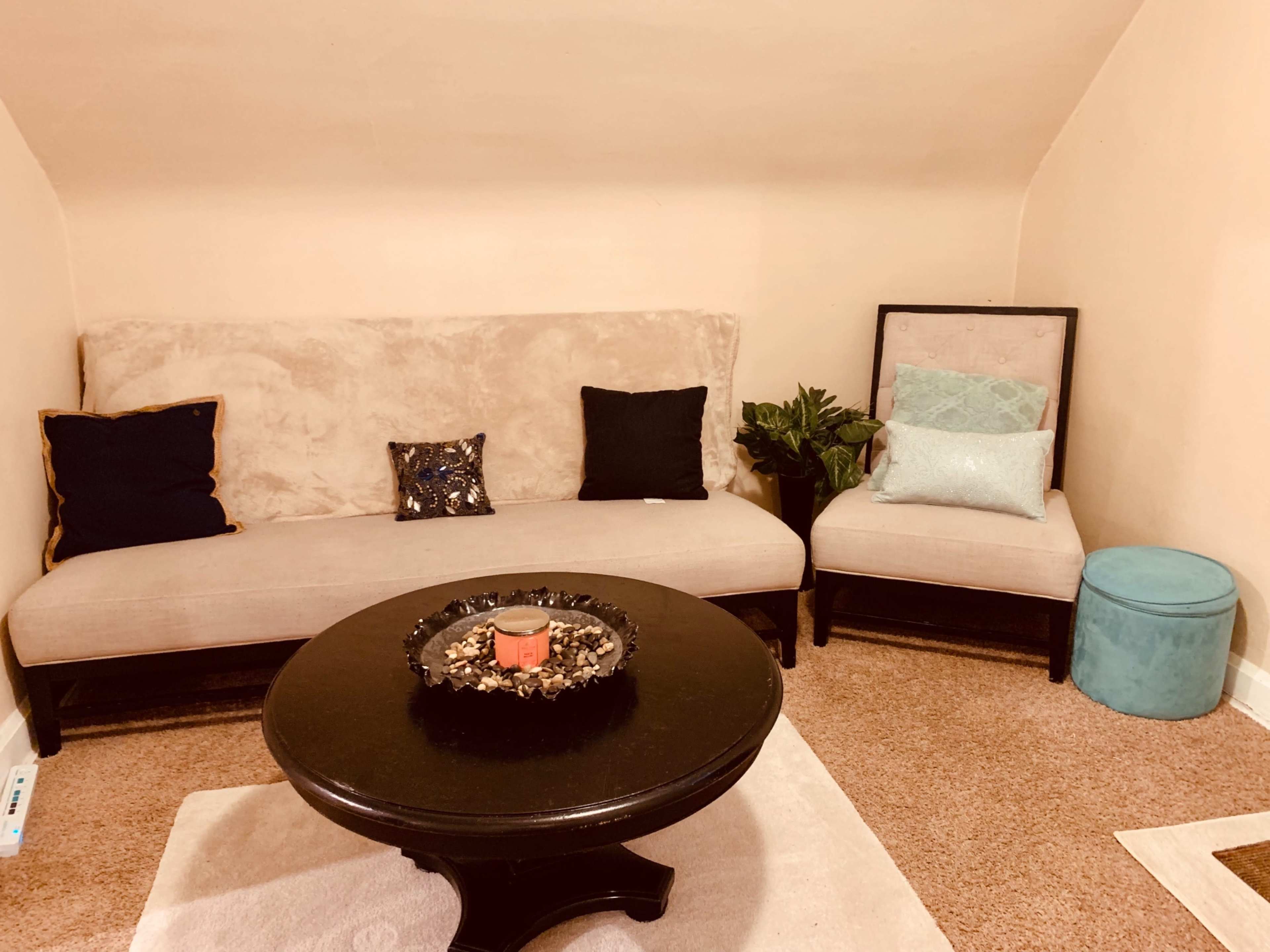 A small room features a beige sofa, an armchair, a round black coffee table with a centerpiece, and a turquoise storage ottoman.