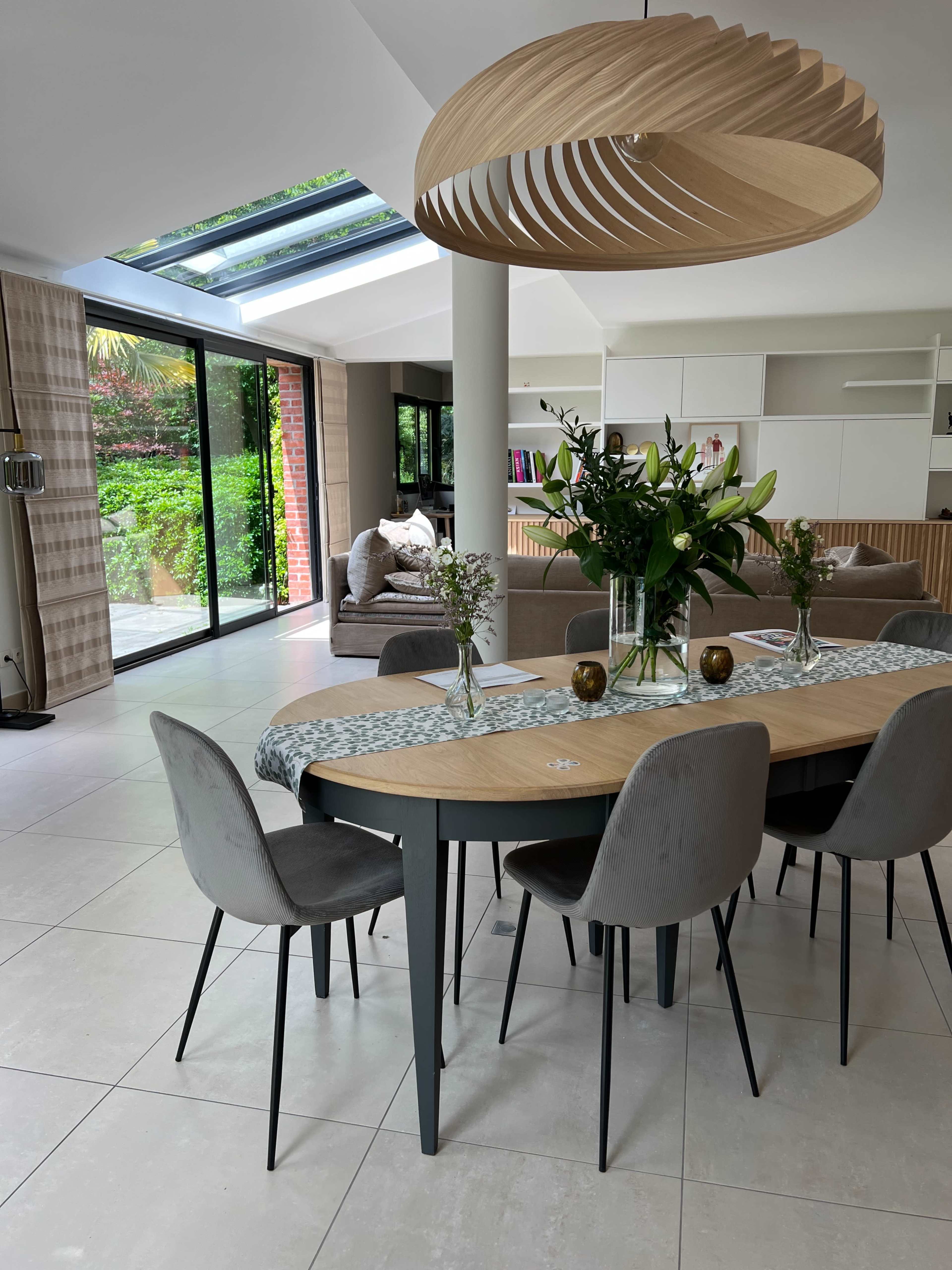 A modern dining area with a wooden table, grey chairs, and a large light fixture, set against a backdrop of large glass windows overlooking a garden.