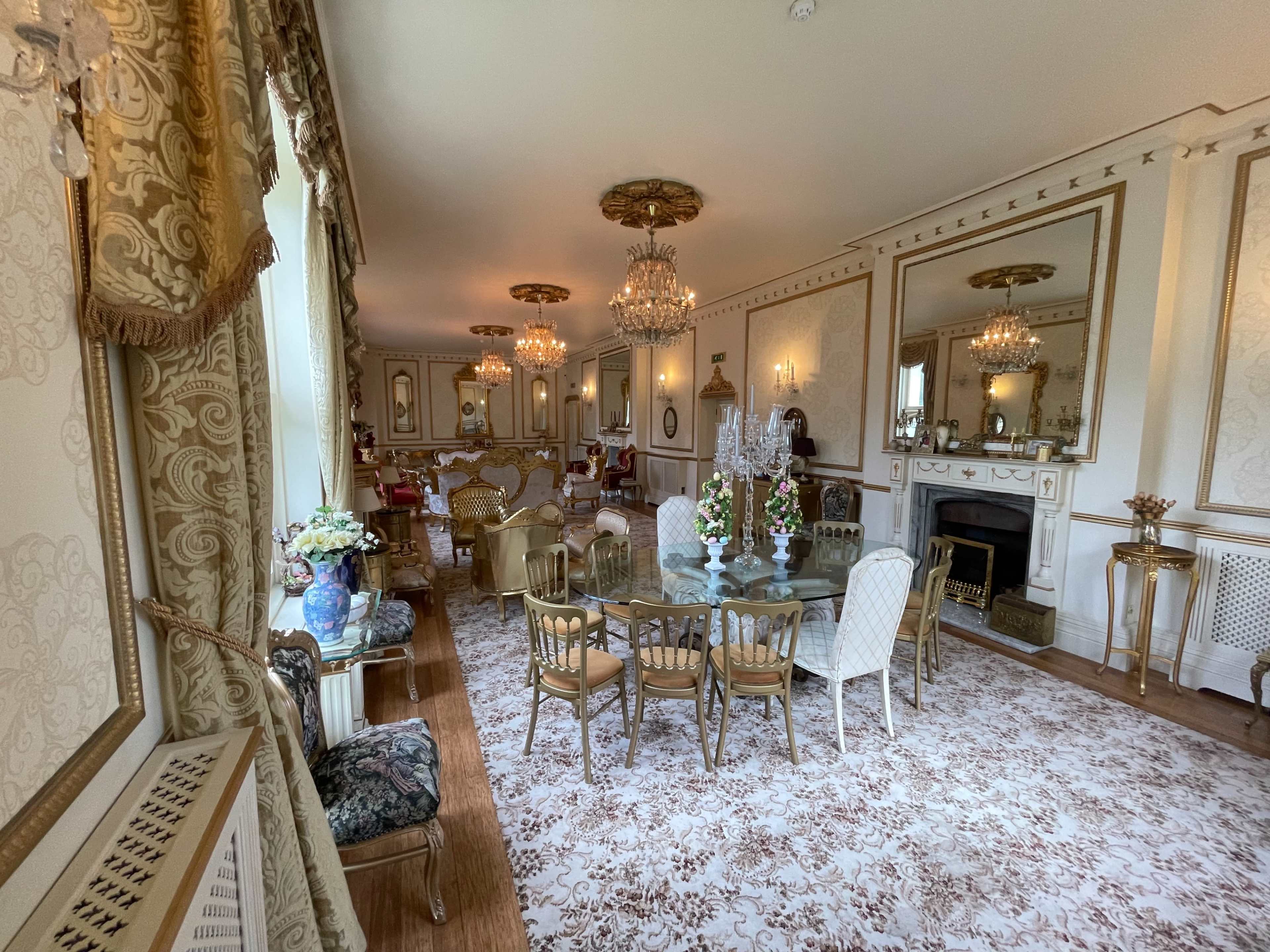 The image shows a spacious, elegantly decorated dining area with a glass table surrounded by ornate chairs, chandeliers hanging from the ceiling, and a large mirror reflecting the room's opulence.