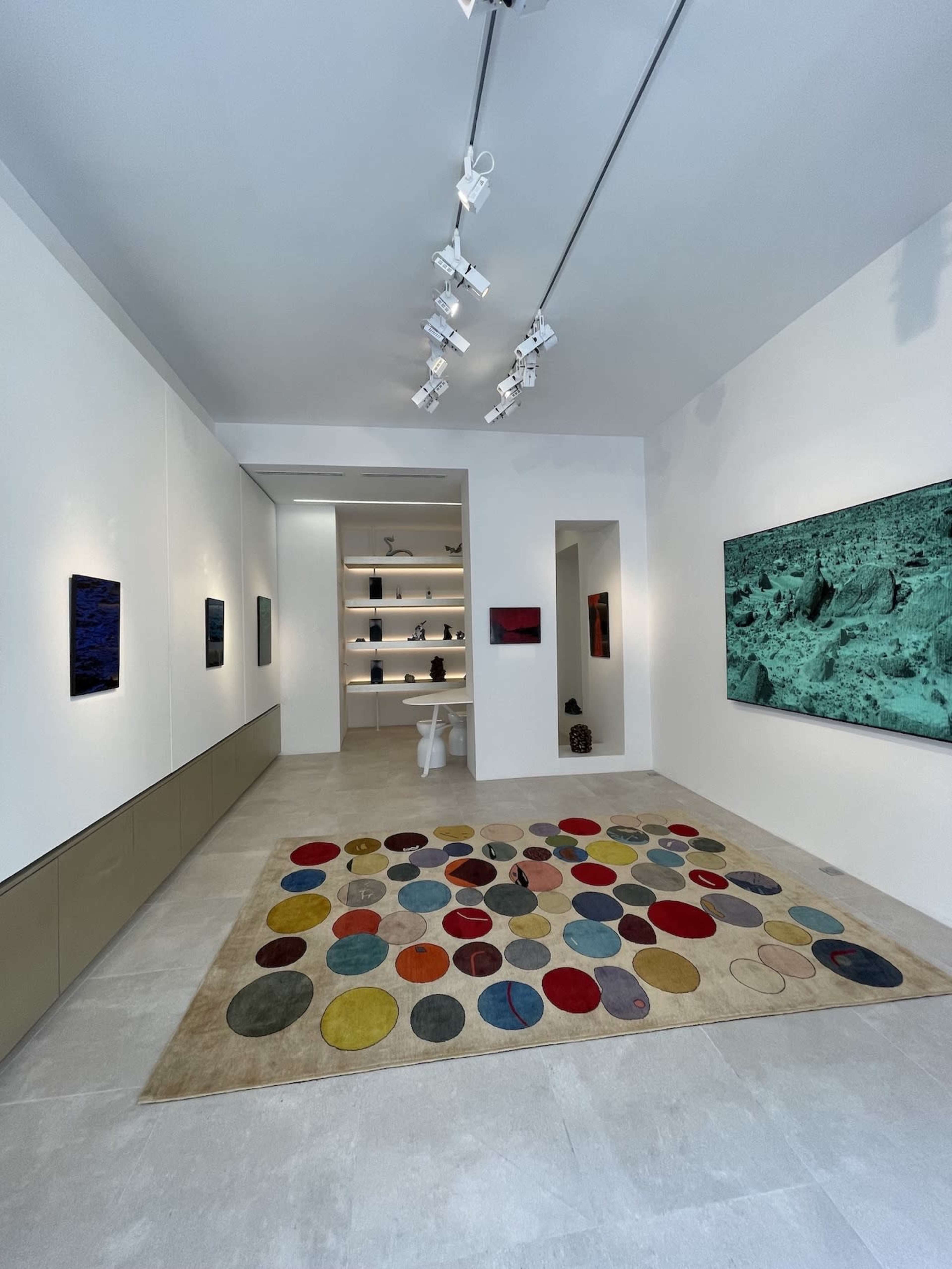 A contemporary art gallery featuring a colorful circular-patterned rug, paintings on the walls, and minimalist displays in a bright, airy space.
