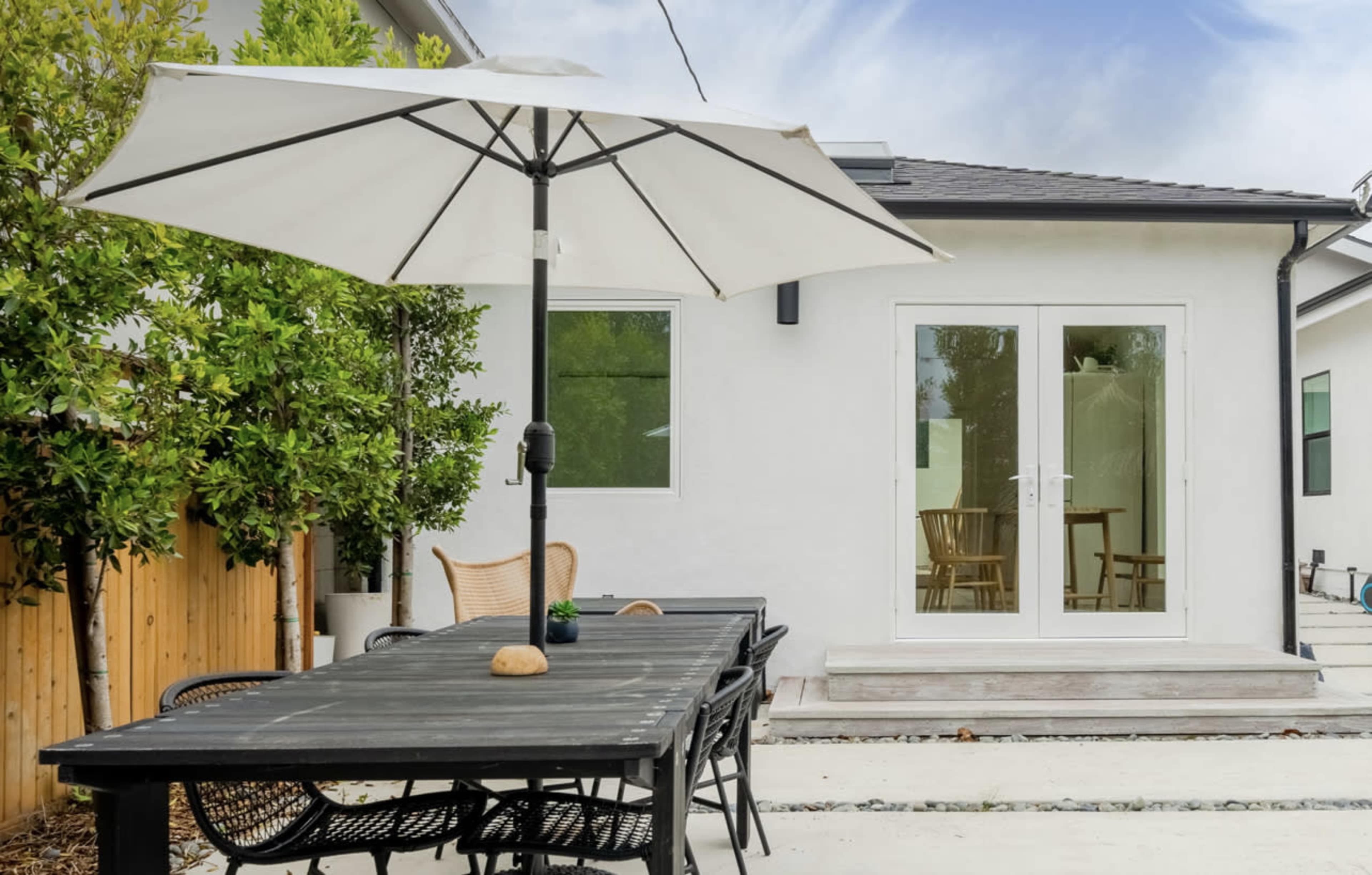 A dining table with black chairs and a white umbrella is set up in an outdoor area beside a building with large glass doors.