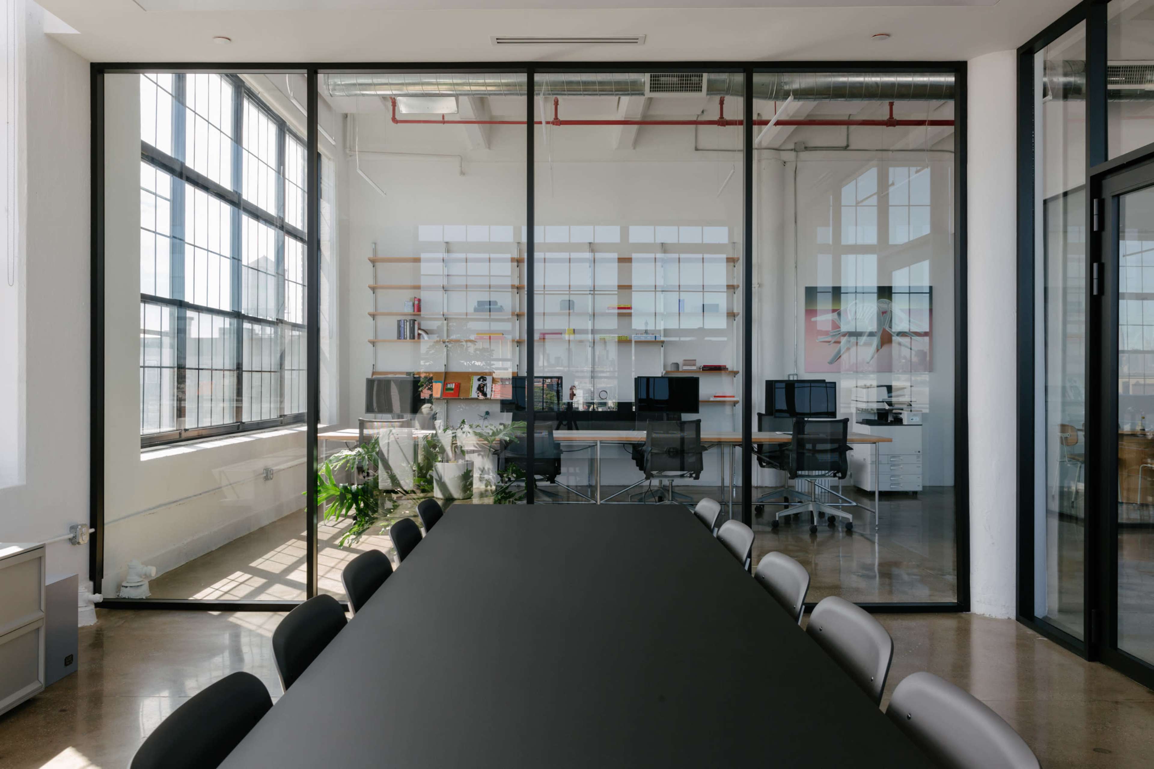 A modern conference room features a long black table and large glass walls that provide a view of a bright office space with desks and shelves.