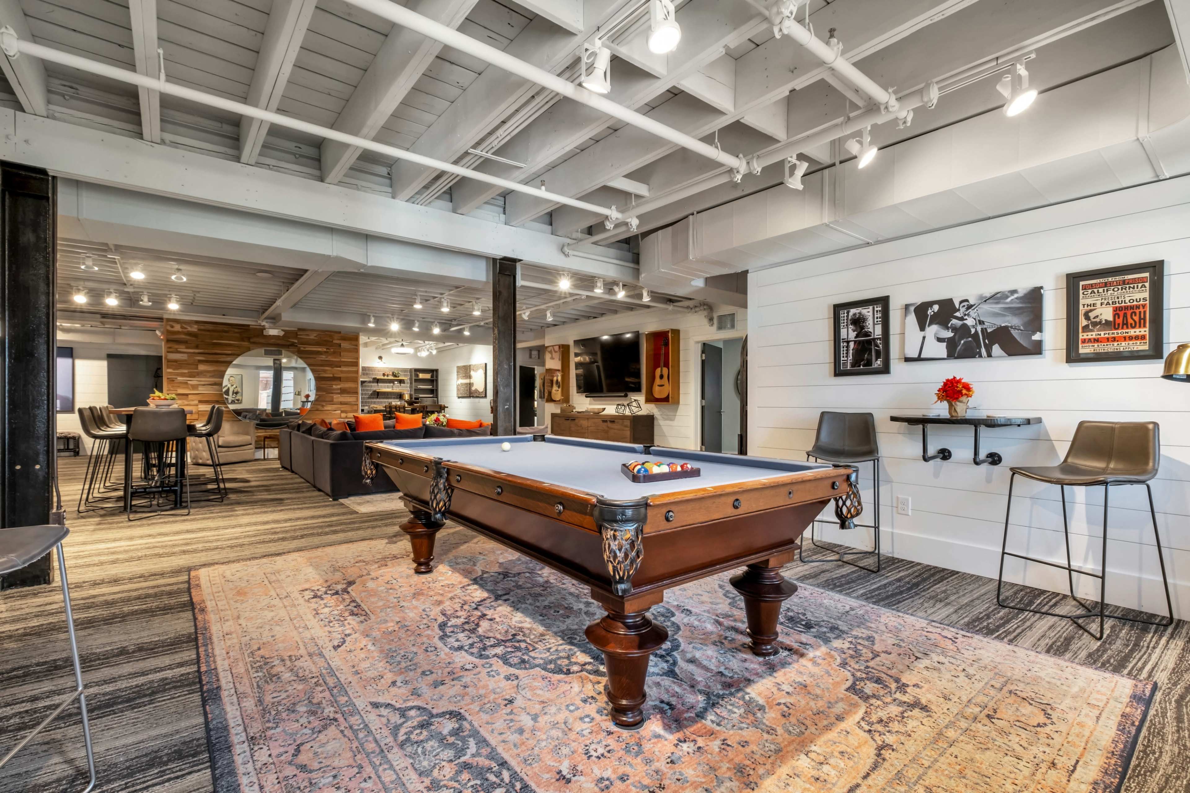 The image shows a well-lit recreational room featuring a billiards table at the center, surrounded by modern furniture and art on the walls.