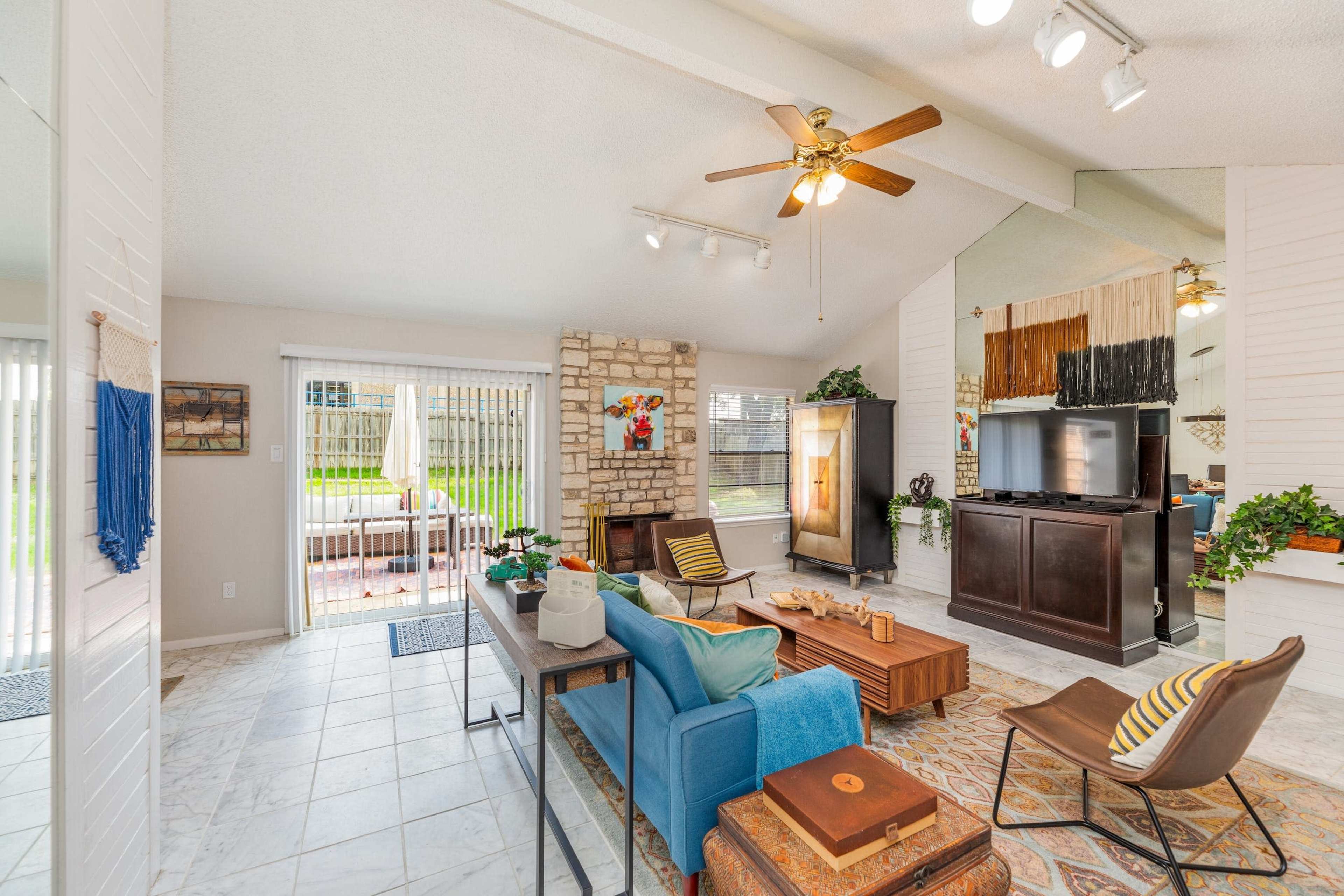 The image shows a spacious living room with a stone fireplace, a large television, and a mix of seating arrangements, all enhanced by natural light from the patio doors.