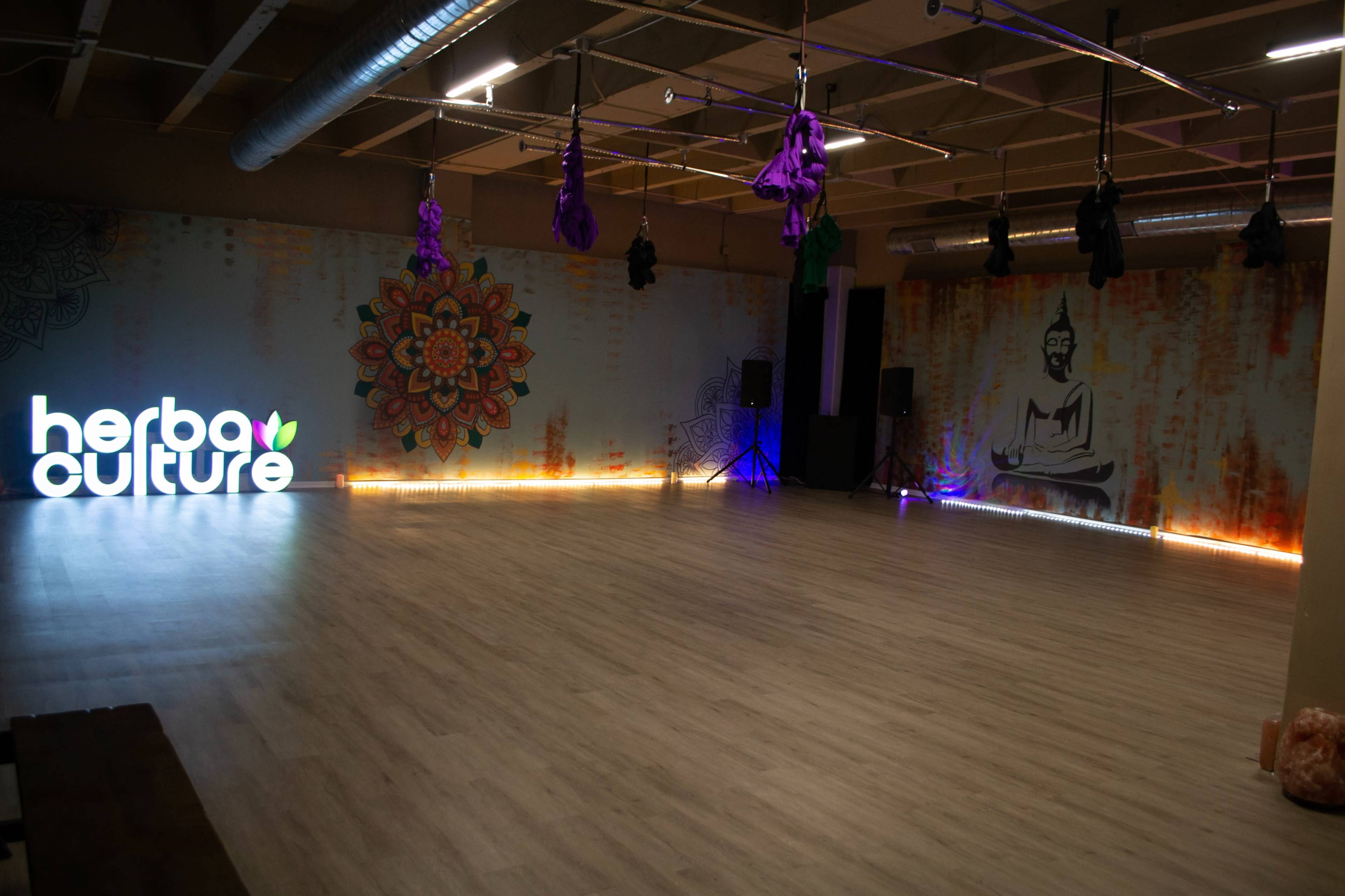 A spacious indoor studio with a hardwood floor, featuring colorful wall artwork, sound equipment, and a brightly lit brand logo for "herba culture."