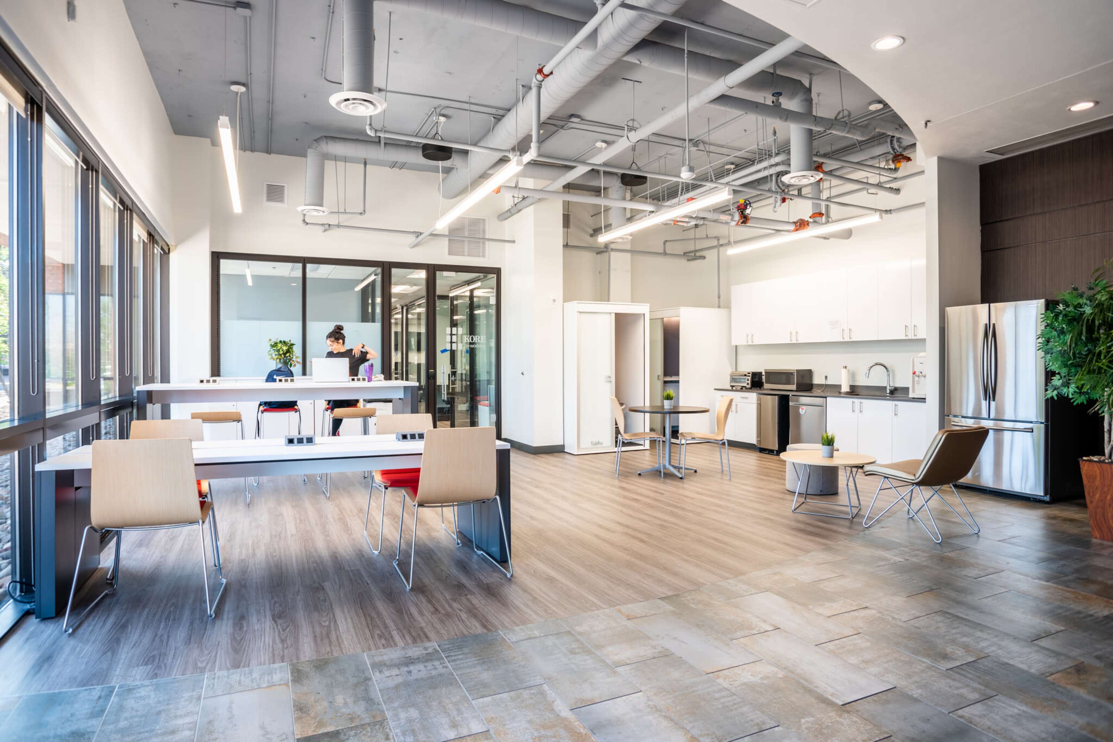 The image shows a modern office kitchen and break area with white cabinets, metal furniture, and large windows allowing natural light.