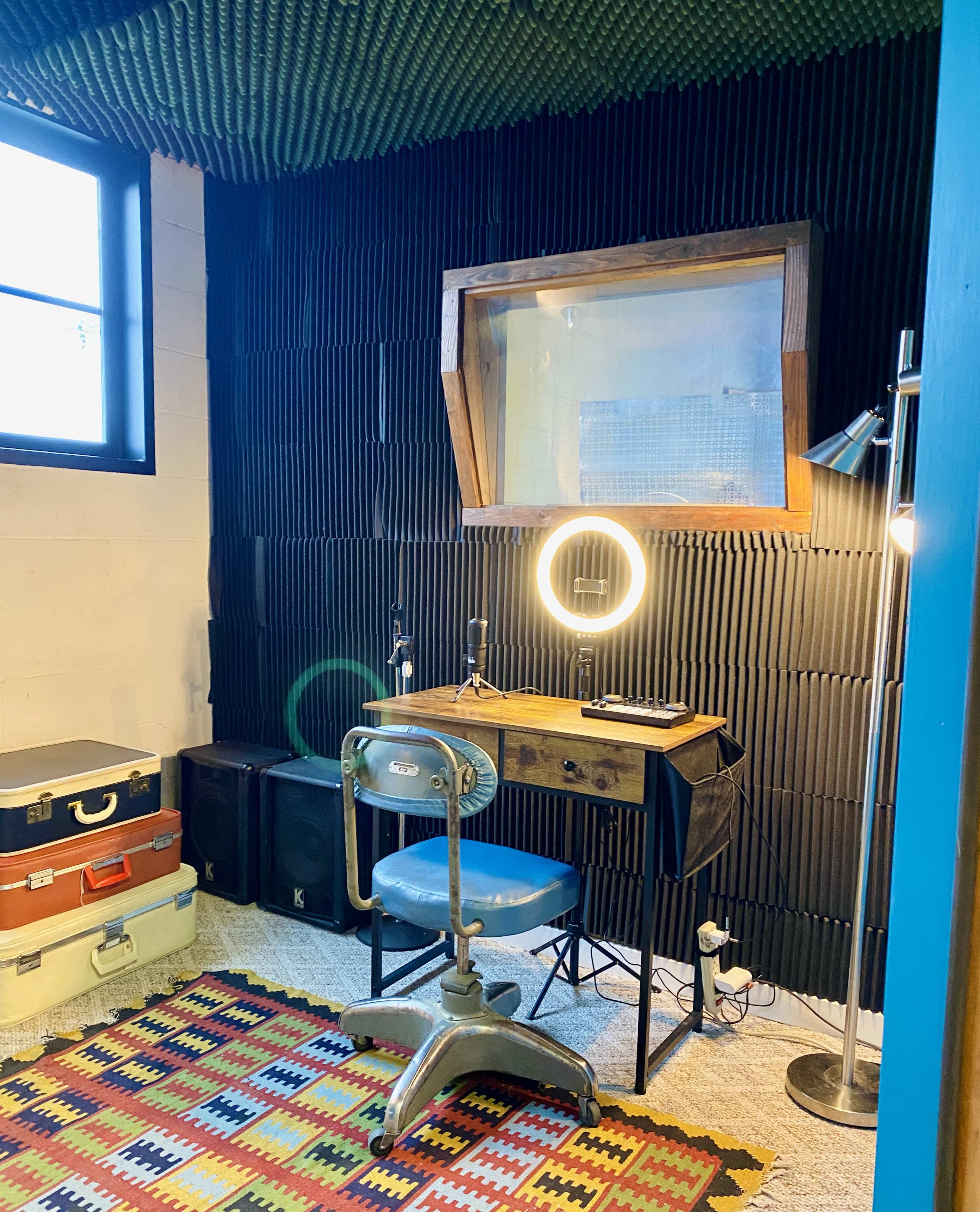 The image shows a compact workspace featuring a desk with a blue chair, a circular mirror light, and various storage boxes, all set against a backdrop of textured black soundproofing panels.