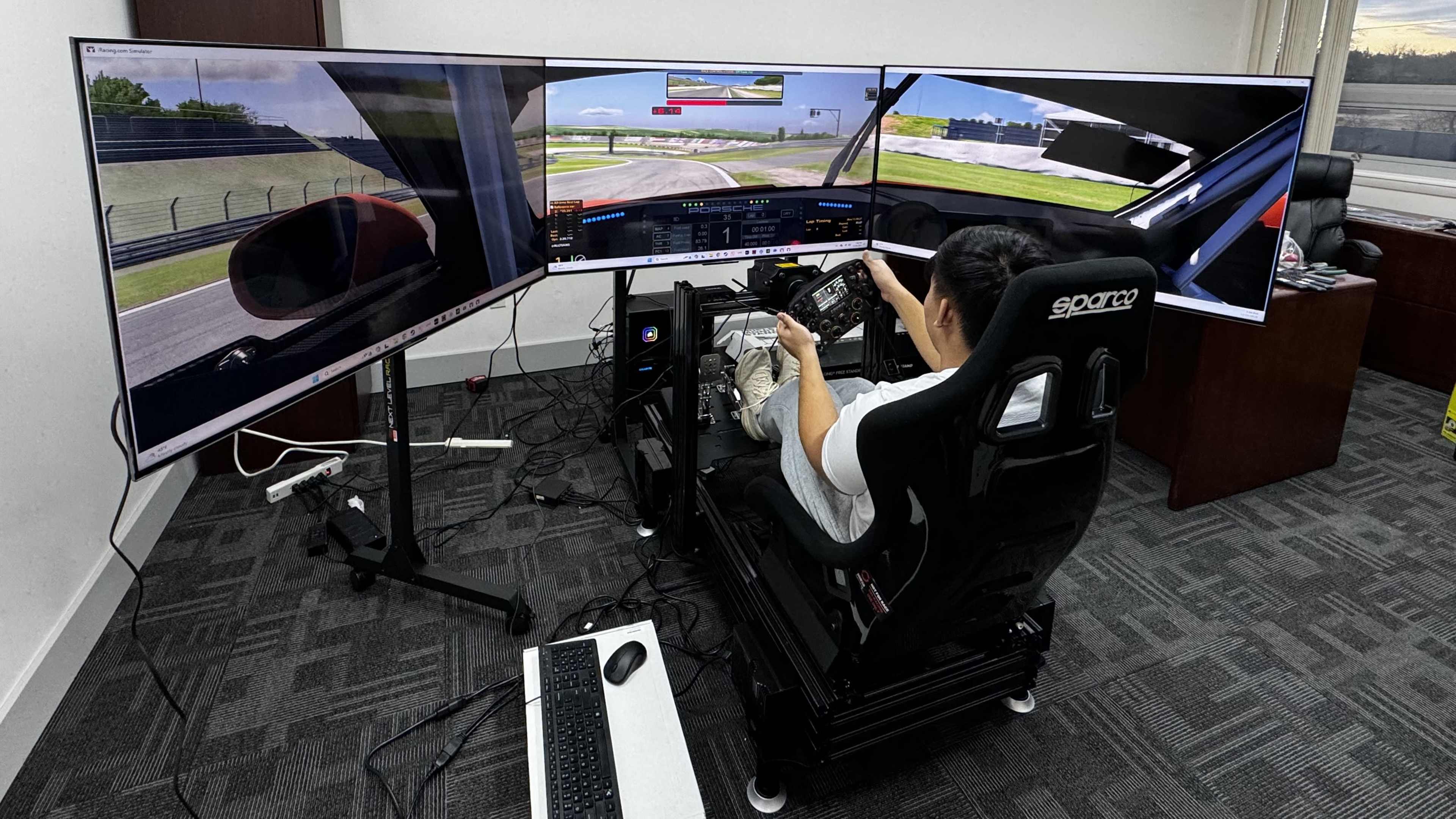 A person sits in a racing simulator setup with three curved monitors displaying a racing track, while using a steering wheel and pedals.