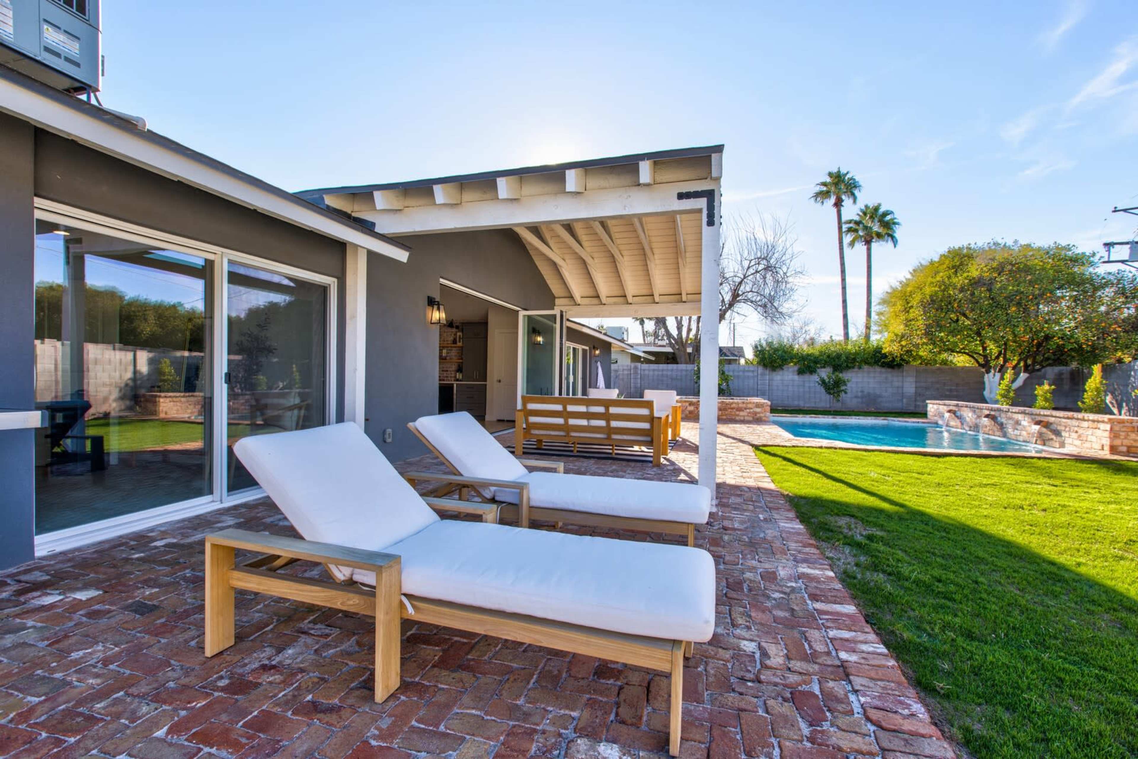 A patio area features two white lounge chairs beside a swimming pool and lush green grass, surrounded by a modern house.