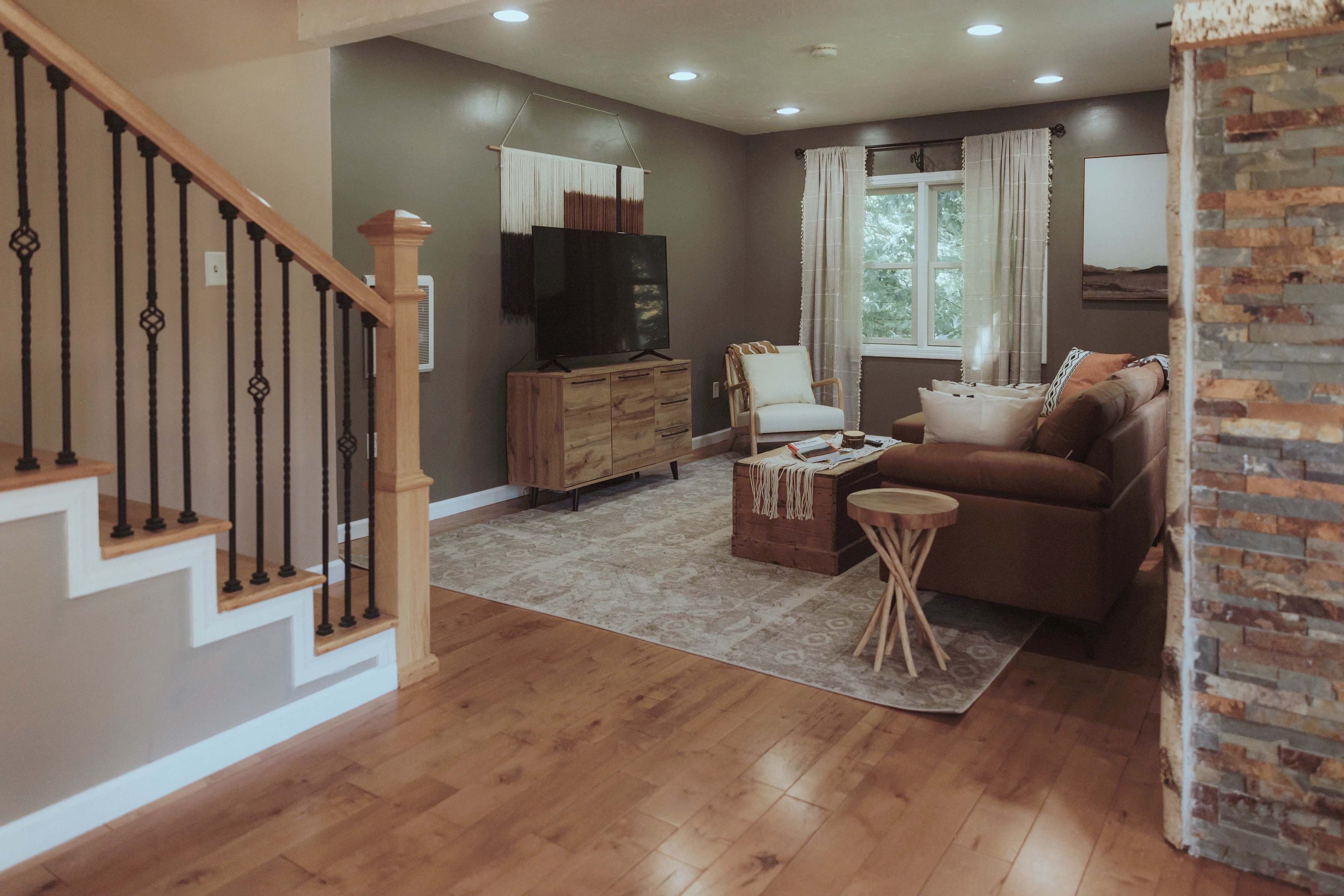 The image shows a cozy living room with a medium brown sofa, a wooden television stand, and large windows dressed with white curtains, alongside a staircase with an iron railing.