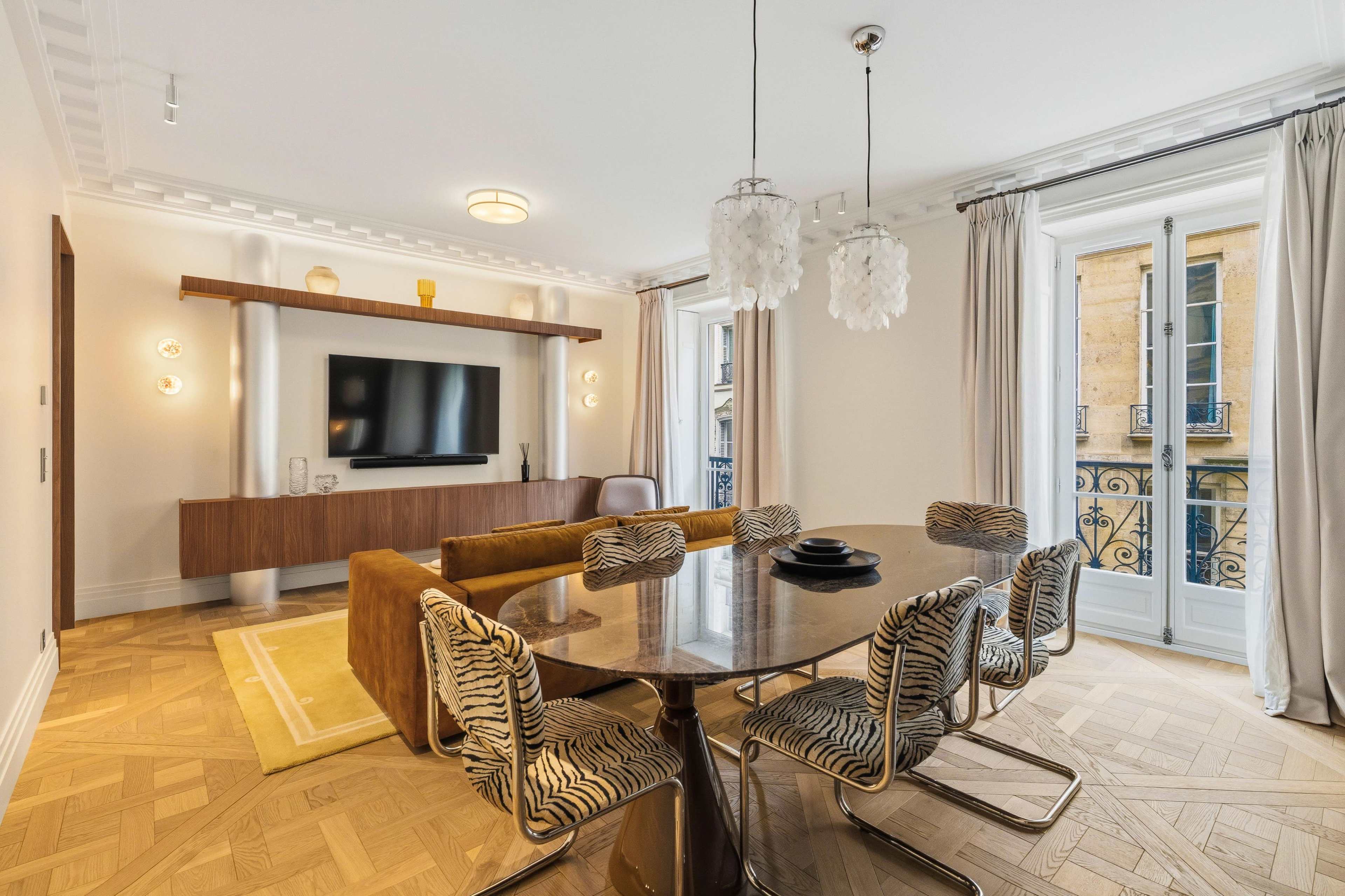 The image shows a modern dining area featuring a round table with patterned chairs, a television mounted on the wall, and large windows with sheer curtains.