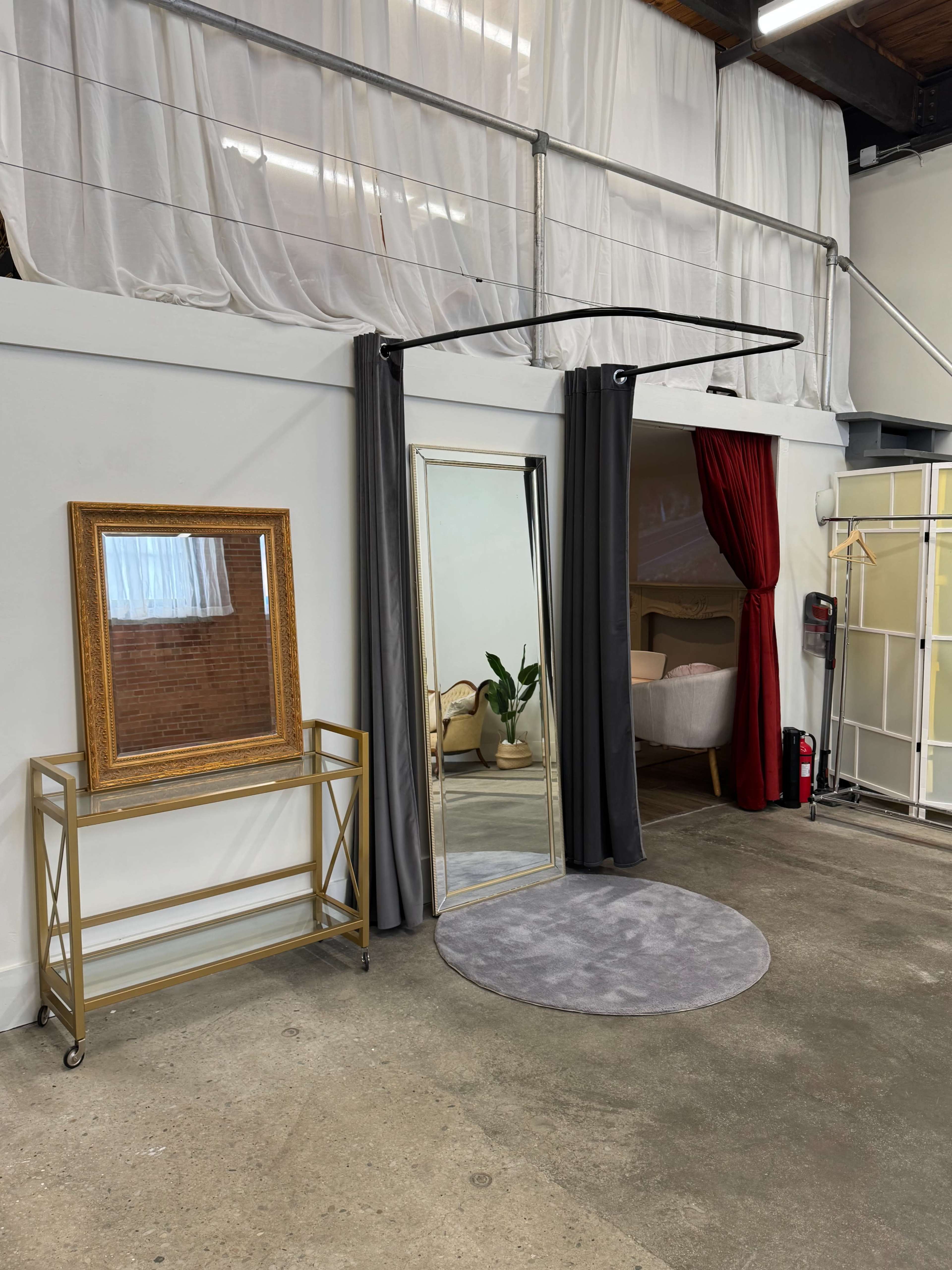 The image shows a stylish interior space featuring a round gray rug, a large mirror framed in gold, a gold cart, and curtains dividing different sections of the room.