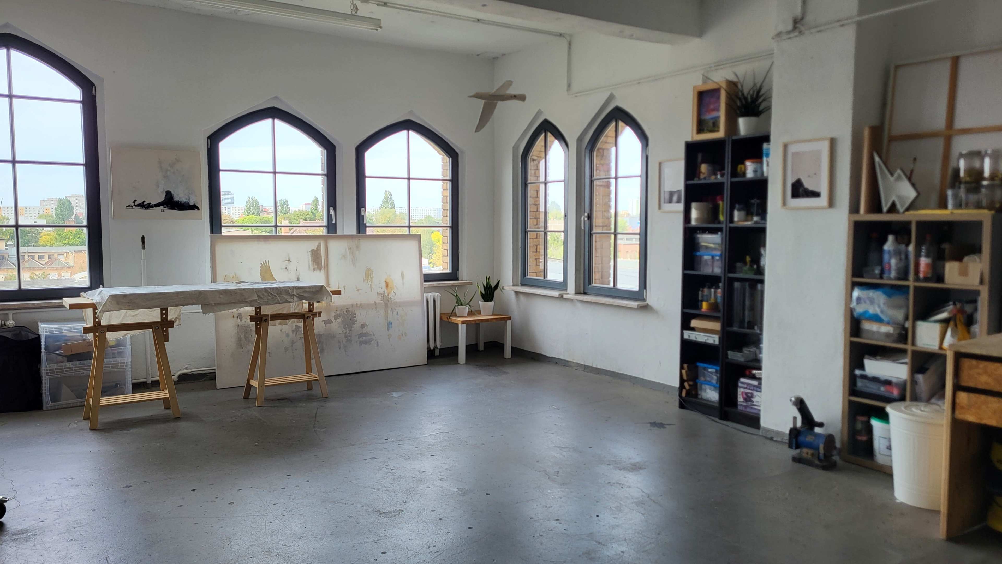 The image shows a spacious, well-lit artist's studio with large arched windows, a worktable, a small table with a plant, and a bookshelf filled with various supplies.