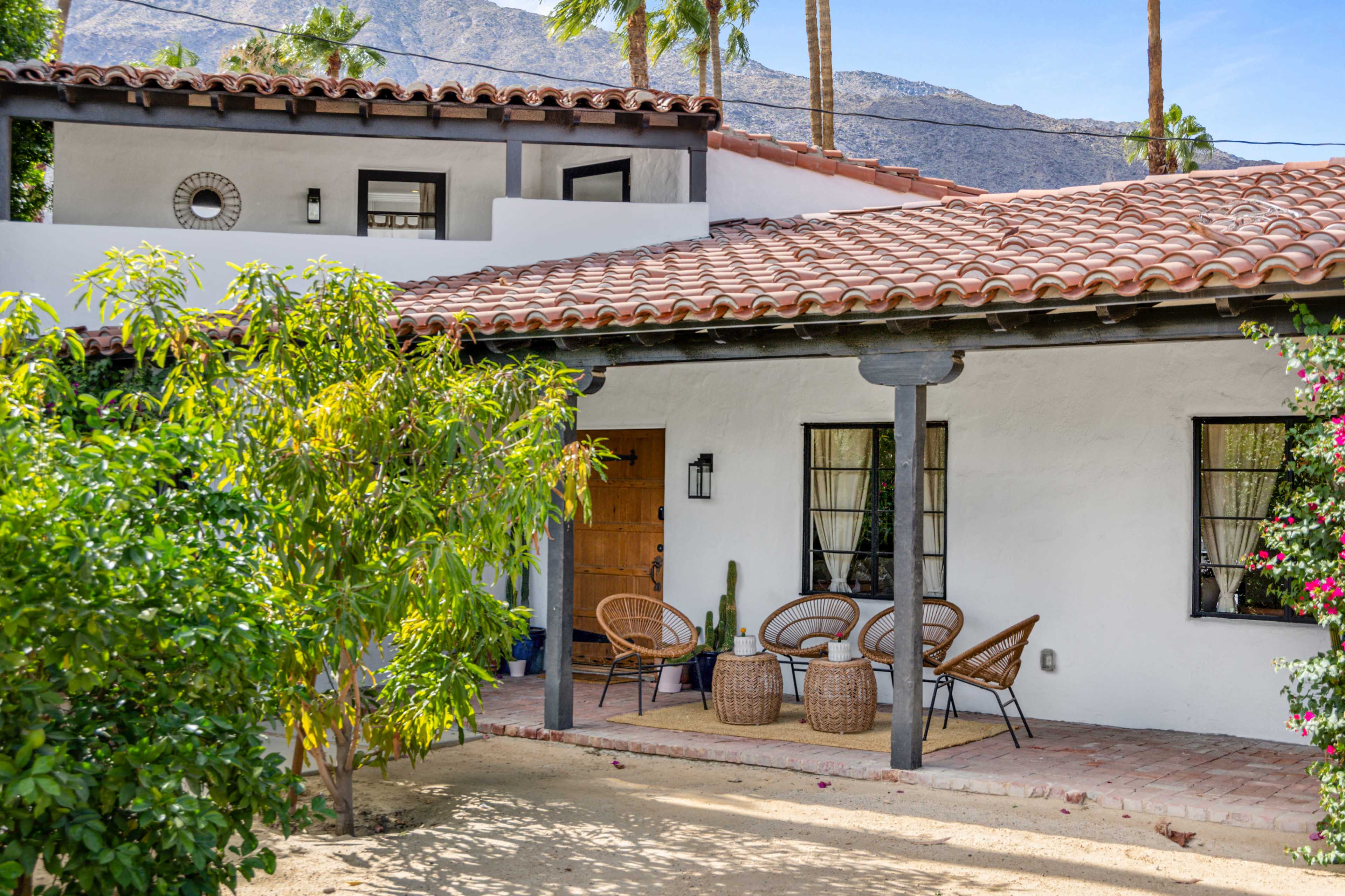 A Spanish-style house with a tiled roof features a porch with wicker chairs and a table, surrounded by greenery and mountains in the background.