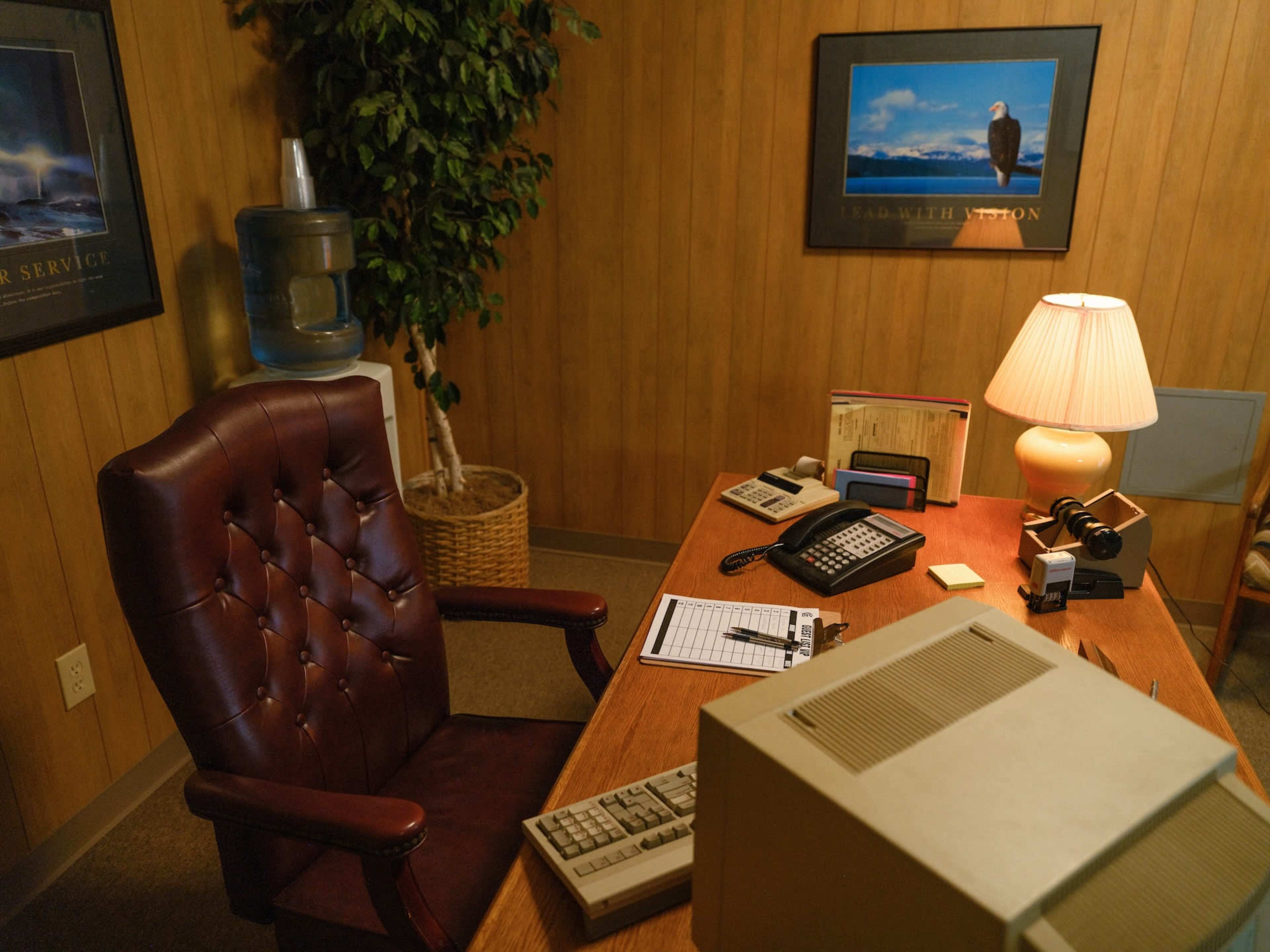 The image shows a small office with a wooden desk, a leather chair, various office equipment, and a poster on the wall featuring an eagle.