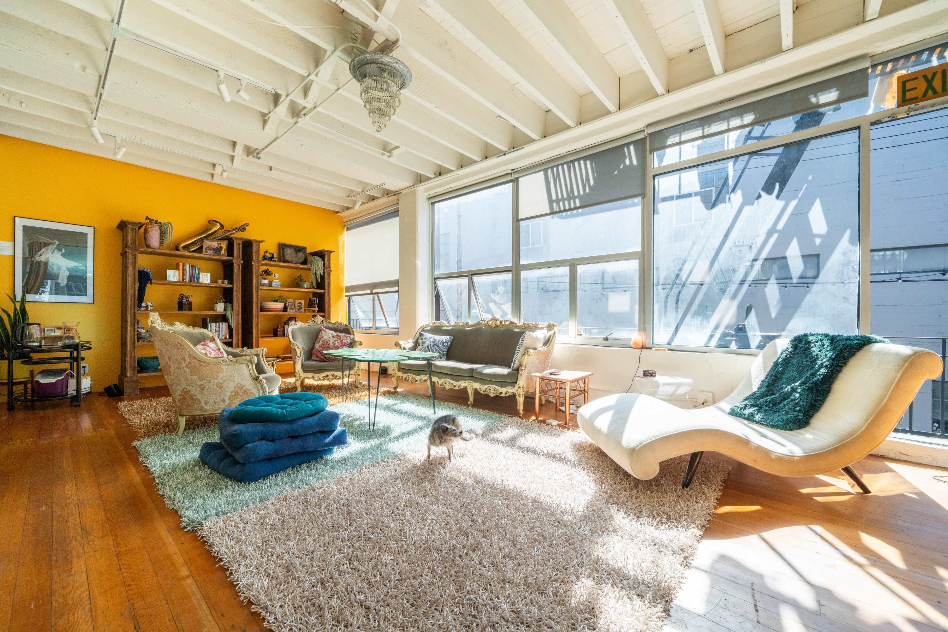 The image shows a spacious living room with wooden floors, large windows, and a cozy seating arrangement that includes a chaise lounge and various soft furnishings.