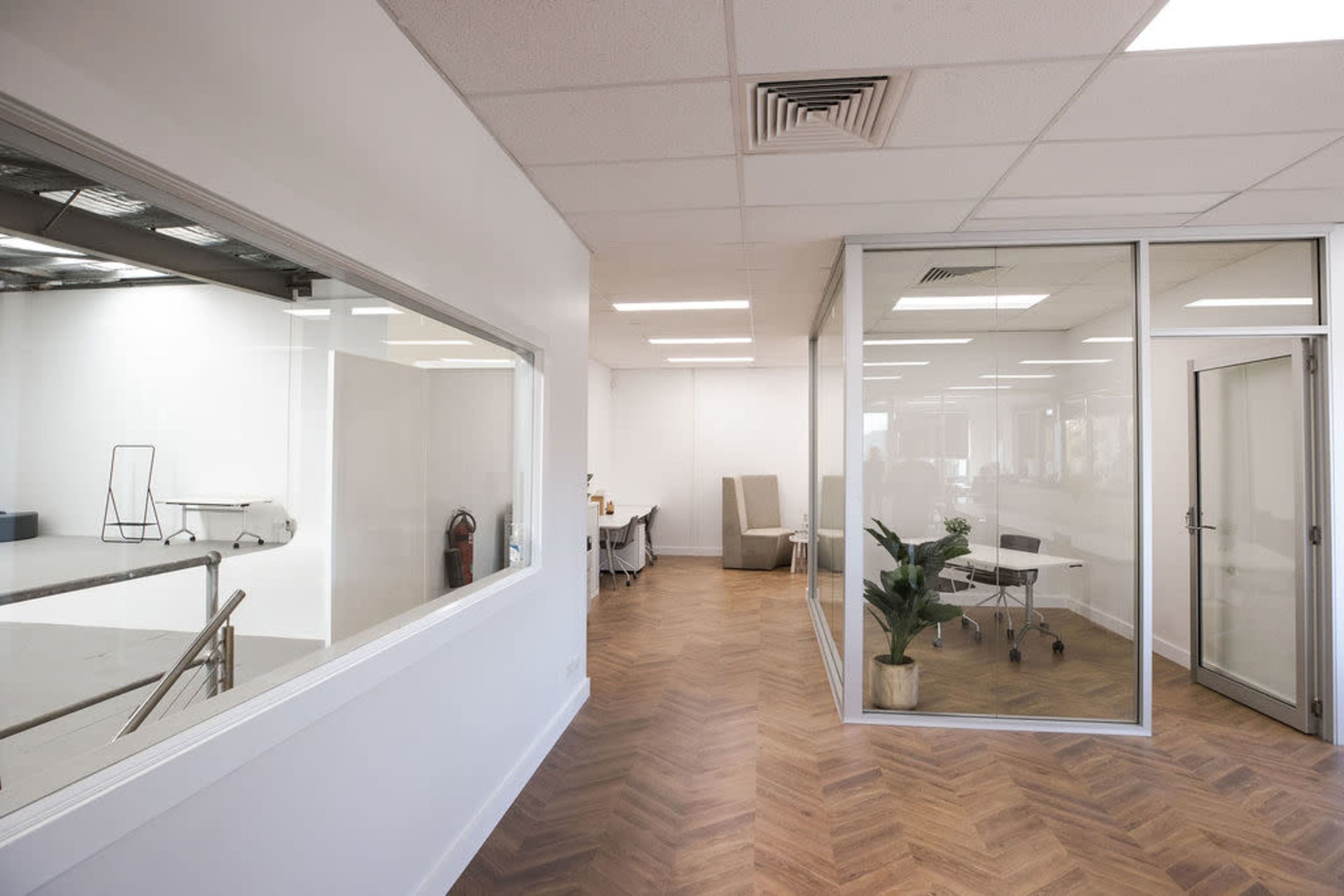 A modern office layout featuring a glass-walled meeting room with a round table and chairs, and an adjacent area with light-colored walls and wooden flooring.