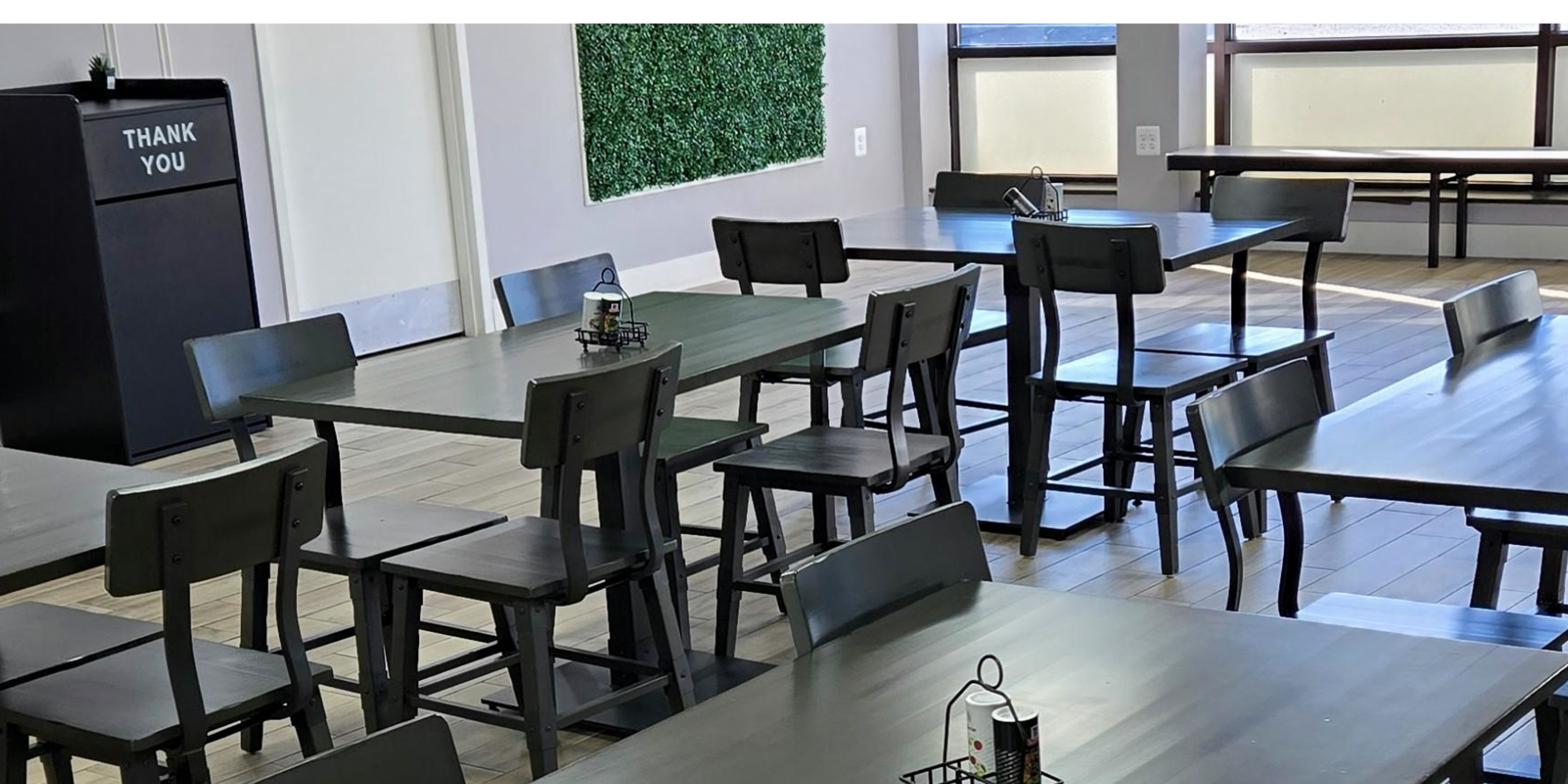 The image shows a dining area with several black wooden tables and chairs arranged neatly, alongside a thank you sign and a decorative green wall feature.