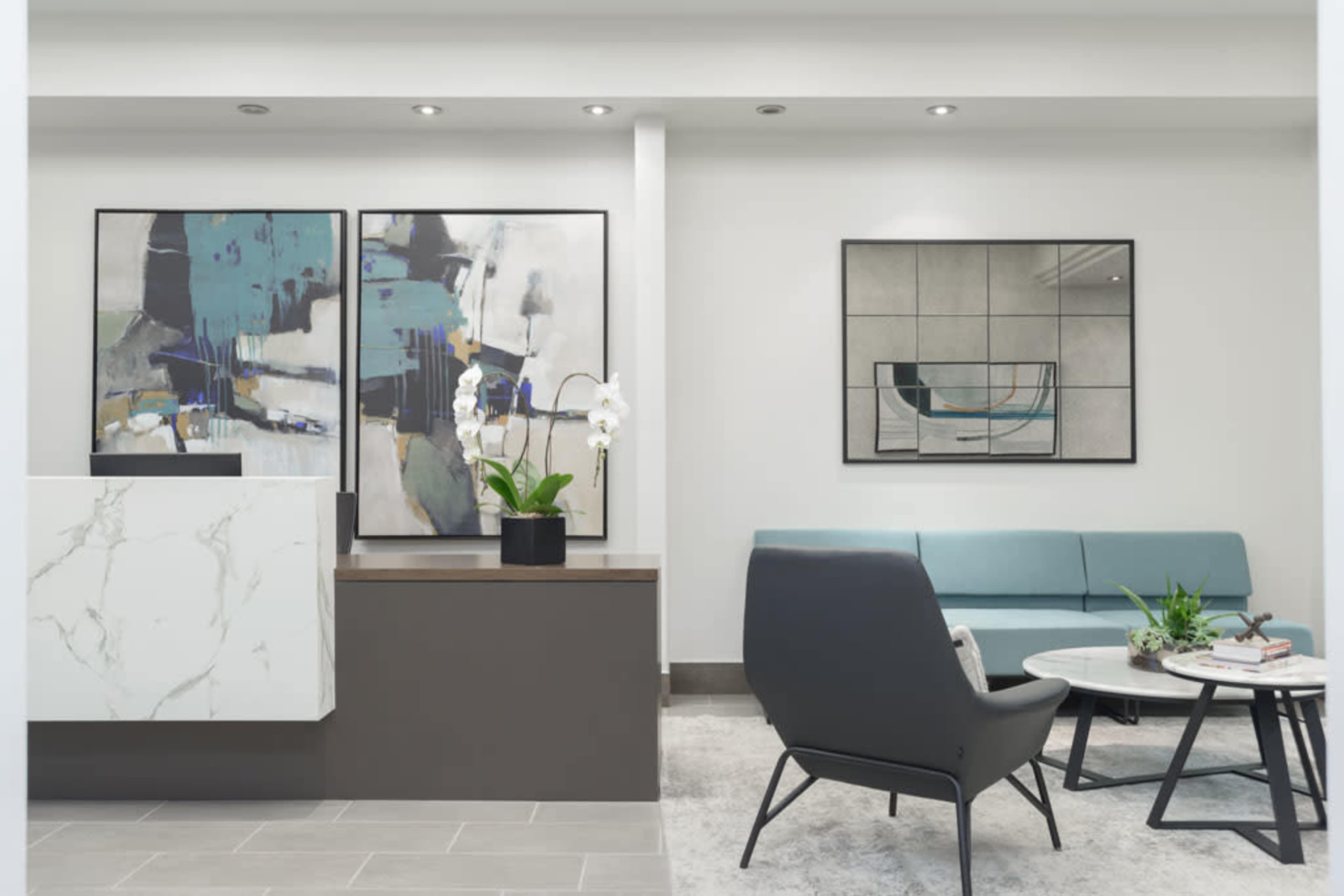 The image shows a modern reception area featuring a marble-fronted desk, two abstract paintings, a light blue sofa, and a round coffee table with plants.