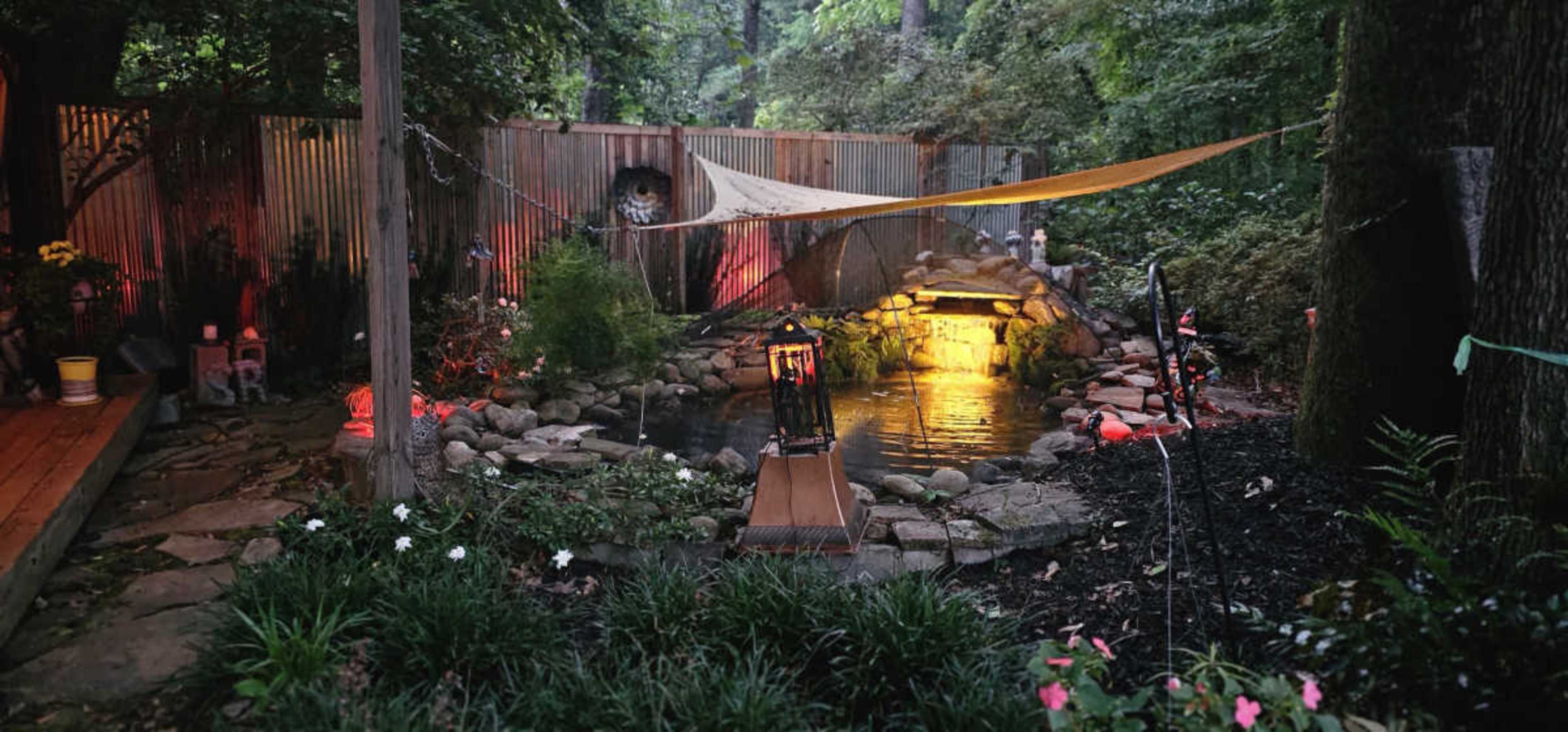 The scene depicts a backyard landscape featuring a pond with a stone fountain, surrounded by lush greenery and illuminated by warm colored lights.