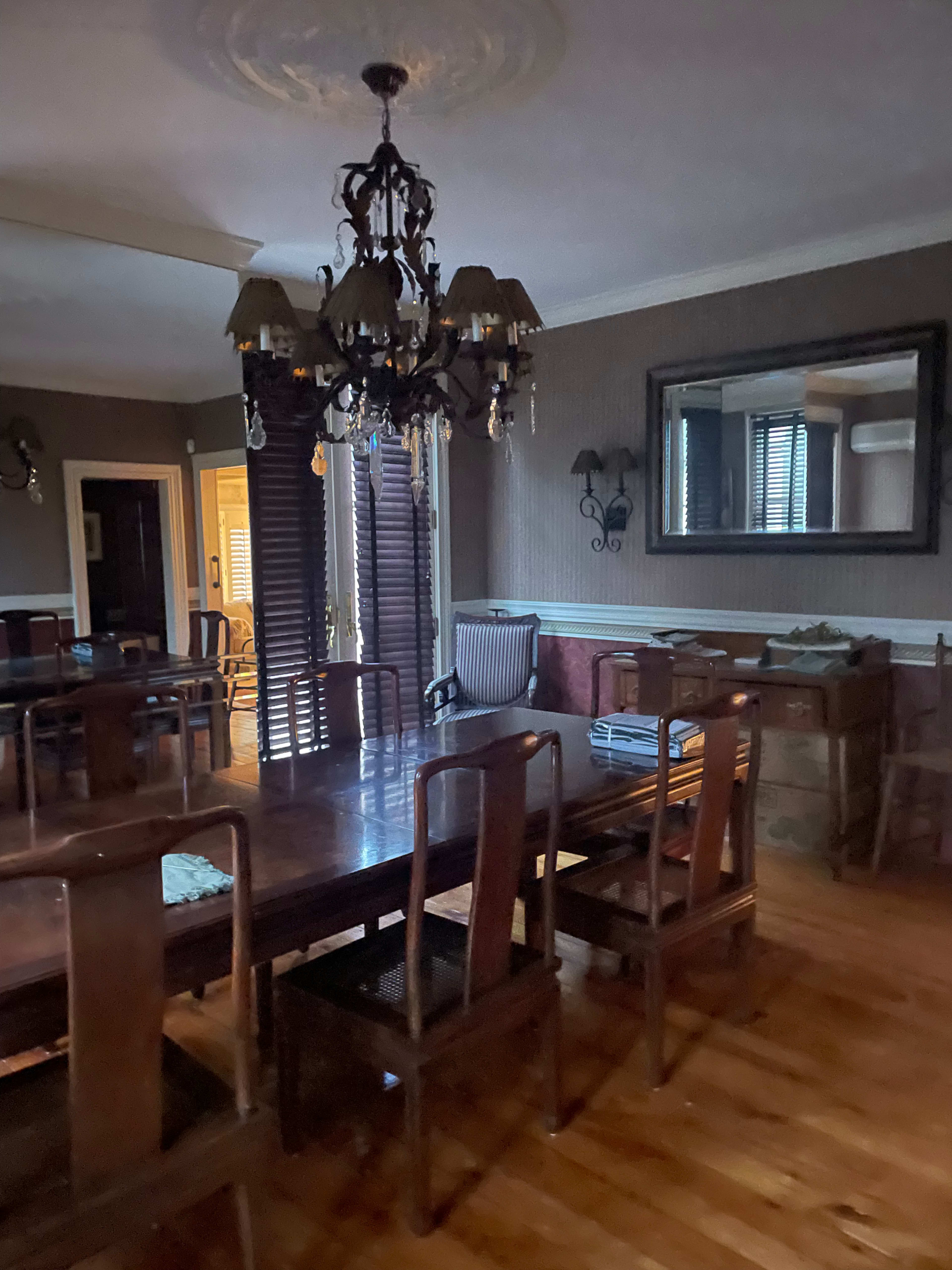 The image shows a dining room featuring a large wooden table surrounded by several chairs, a chandelier hanging from the ceiling, and a mirror reflecting the room's decor.
