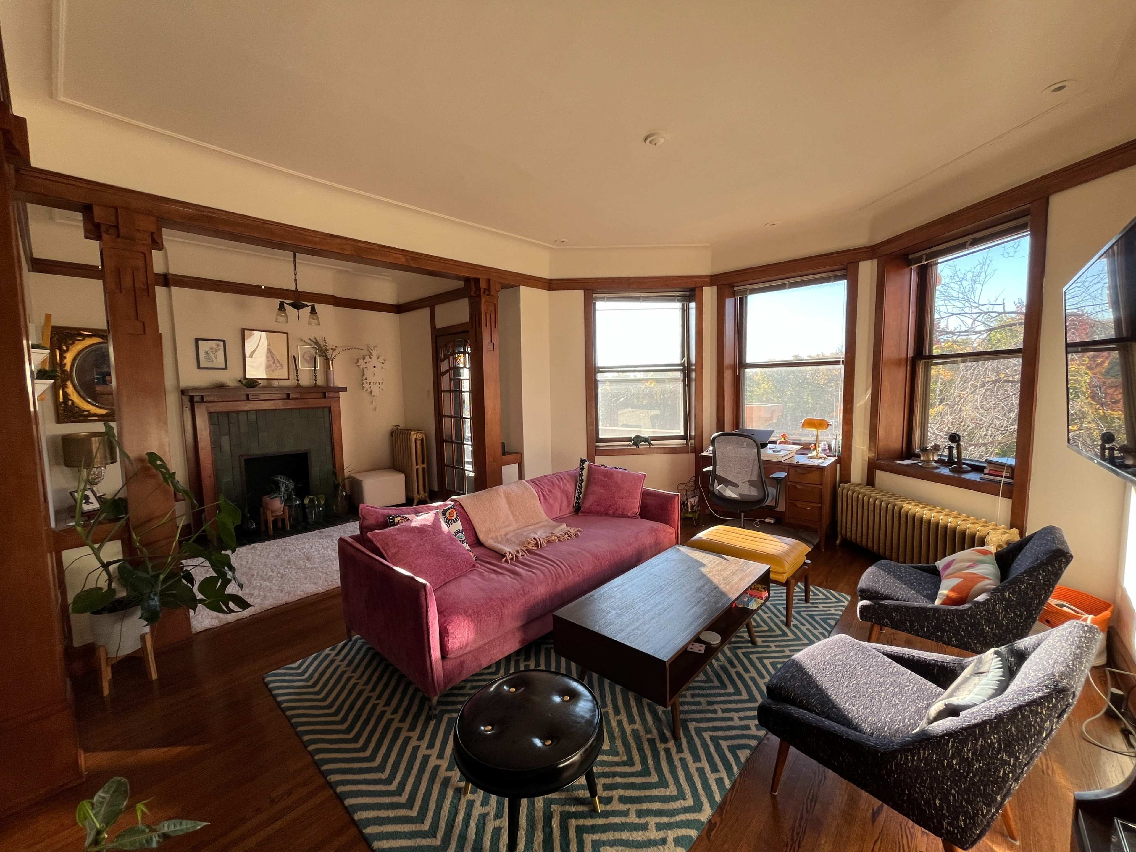 The image shows a cozy living room with a pink couch, wooden furniture, and large windows that provide natural light.