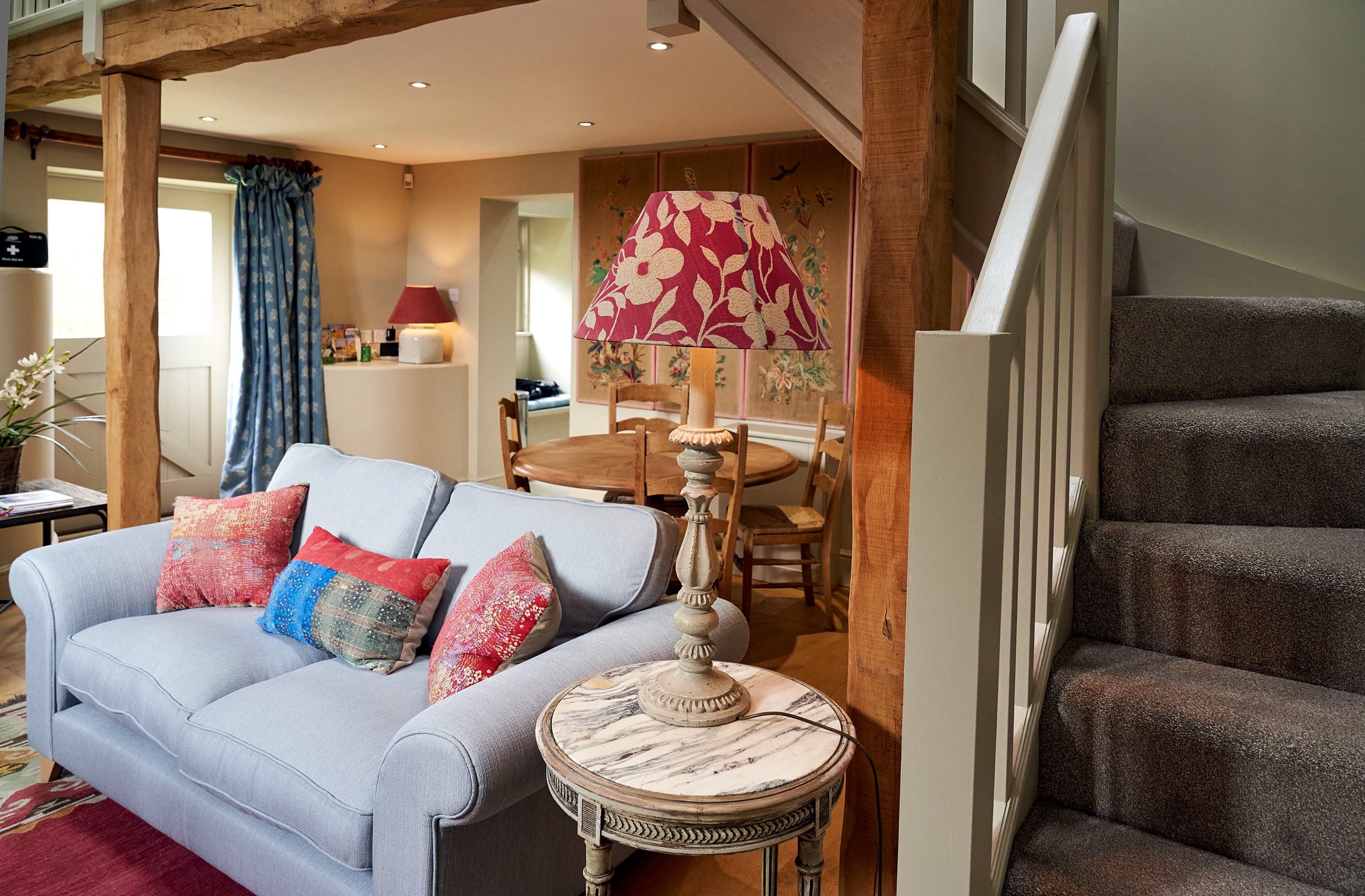 A cozy living area features a light blue sofa with patterned cushions, a wooden side table with a decorative lamp, and a staircase leading to an upper level.