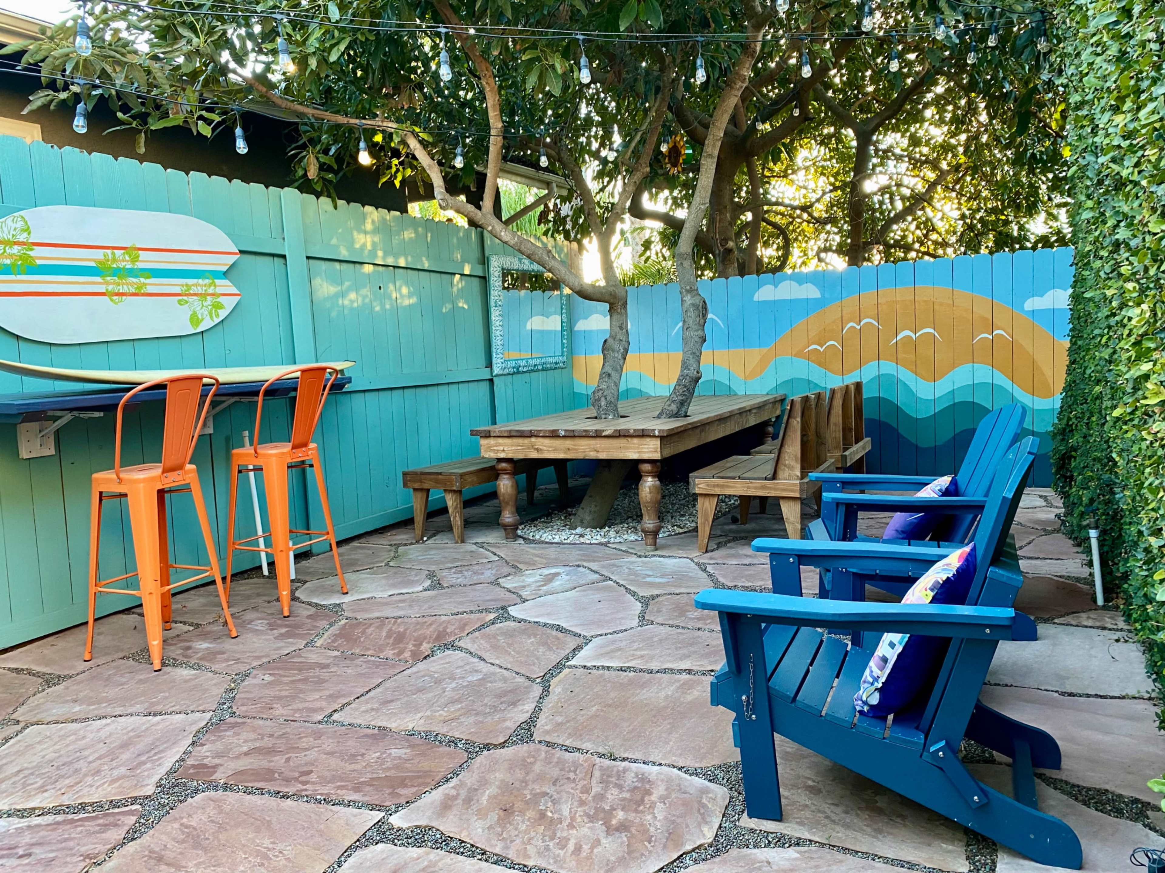 A patio area with a wooden table, colorful chairs, and a painted mural on the fence depicting mountains and a surfboard.