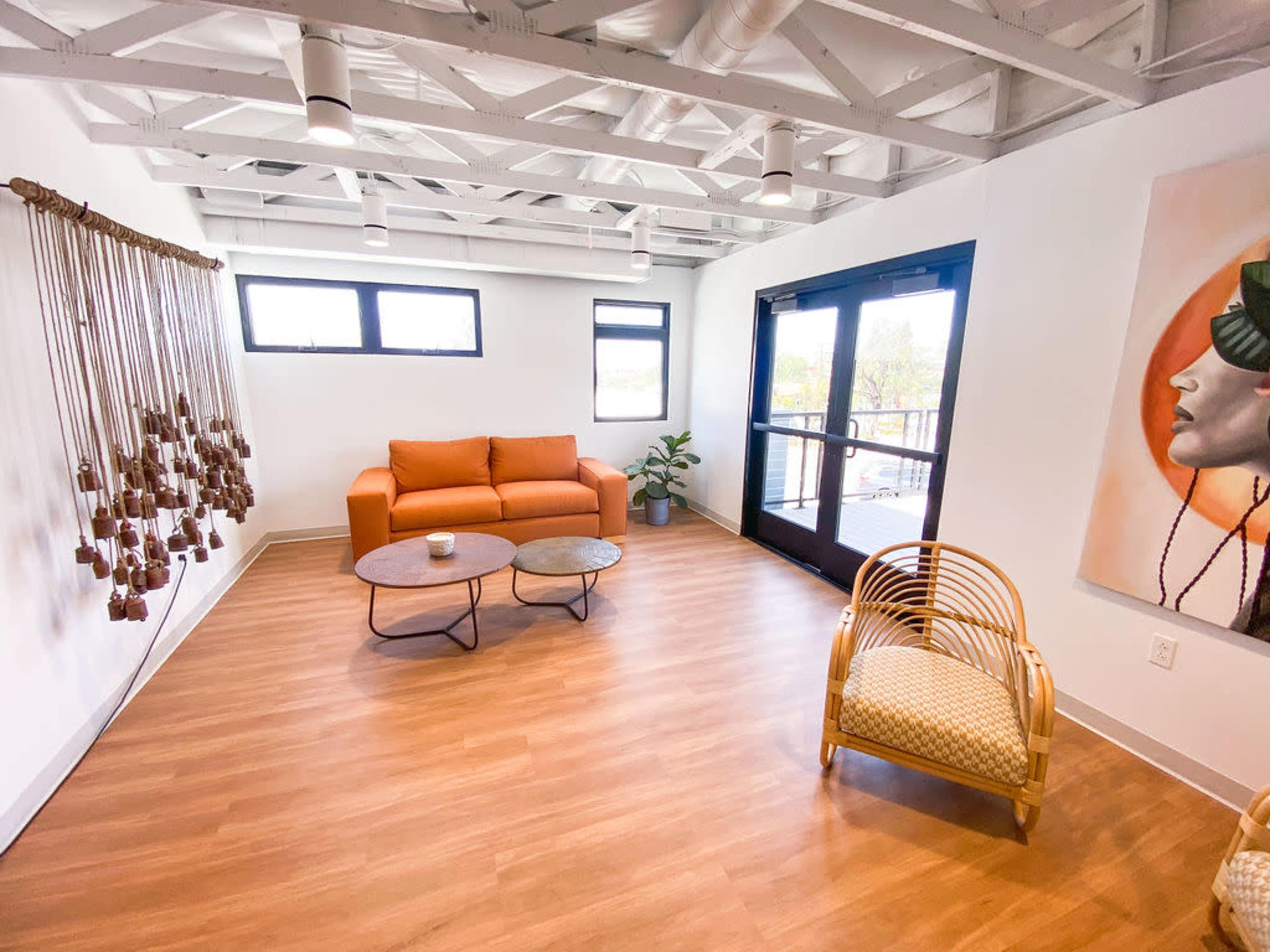 A modern interior space features a brown couch, a round coffee table, and a wicker chair, with large windows and wooden flooring.