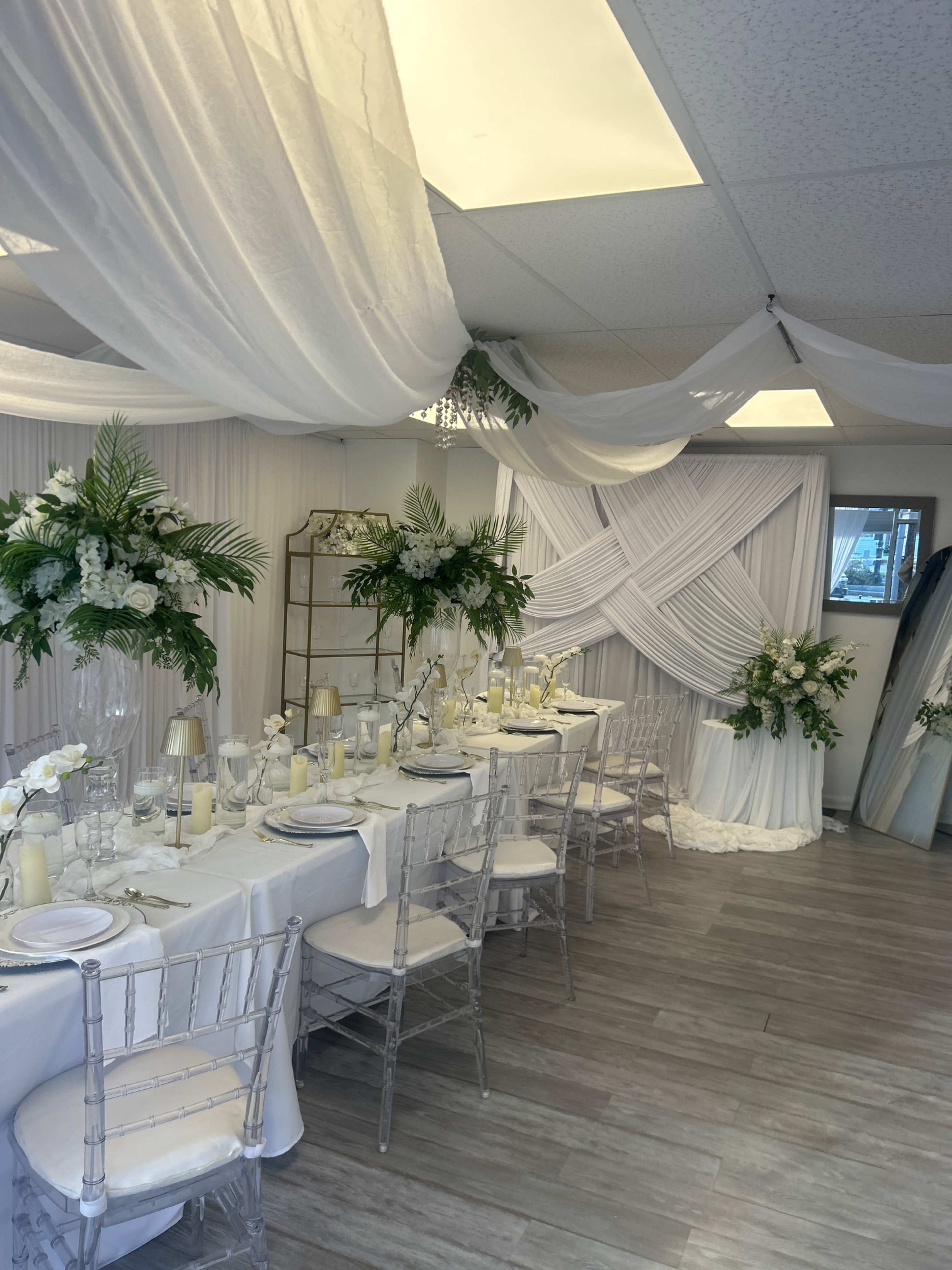 The image depicts a decorated event space with white drapery, elegant table settings, and floral arrangements.