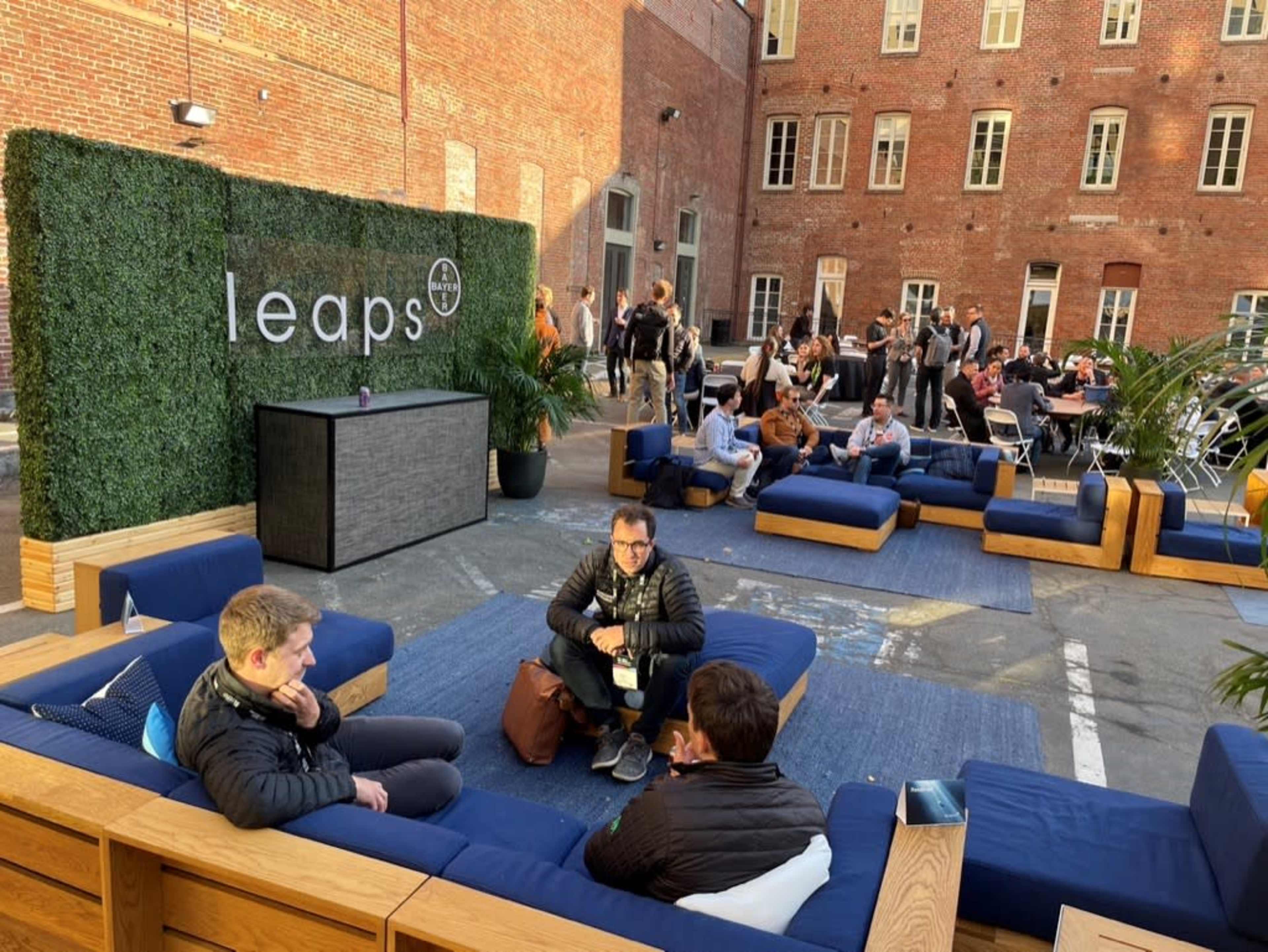 A courtyard features modern seating arrangements with attendees engaged in conversation, surrounded by greenery and a backdrop of brick buildings.