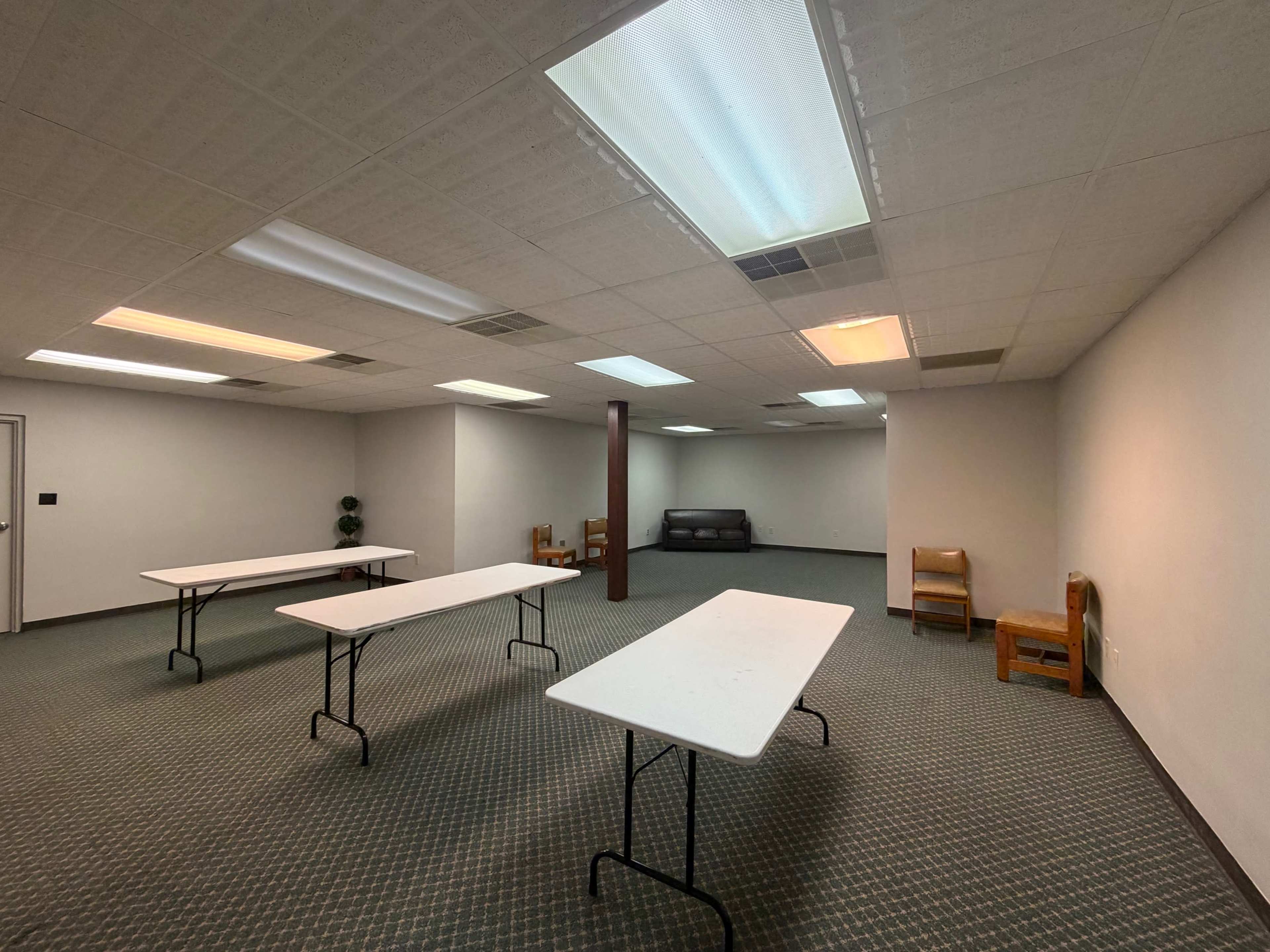 The image shows a sparsely furnished interior room with four white tables, several chairs, and a couch against the wall.