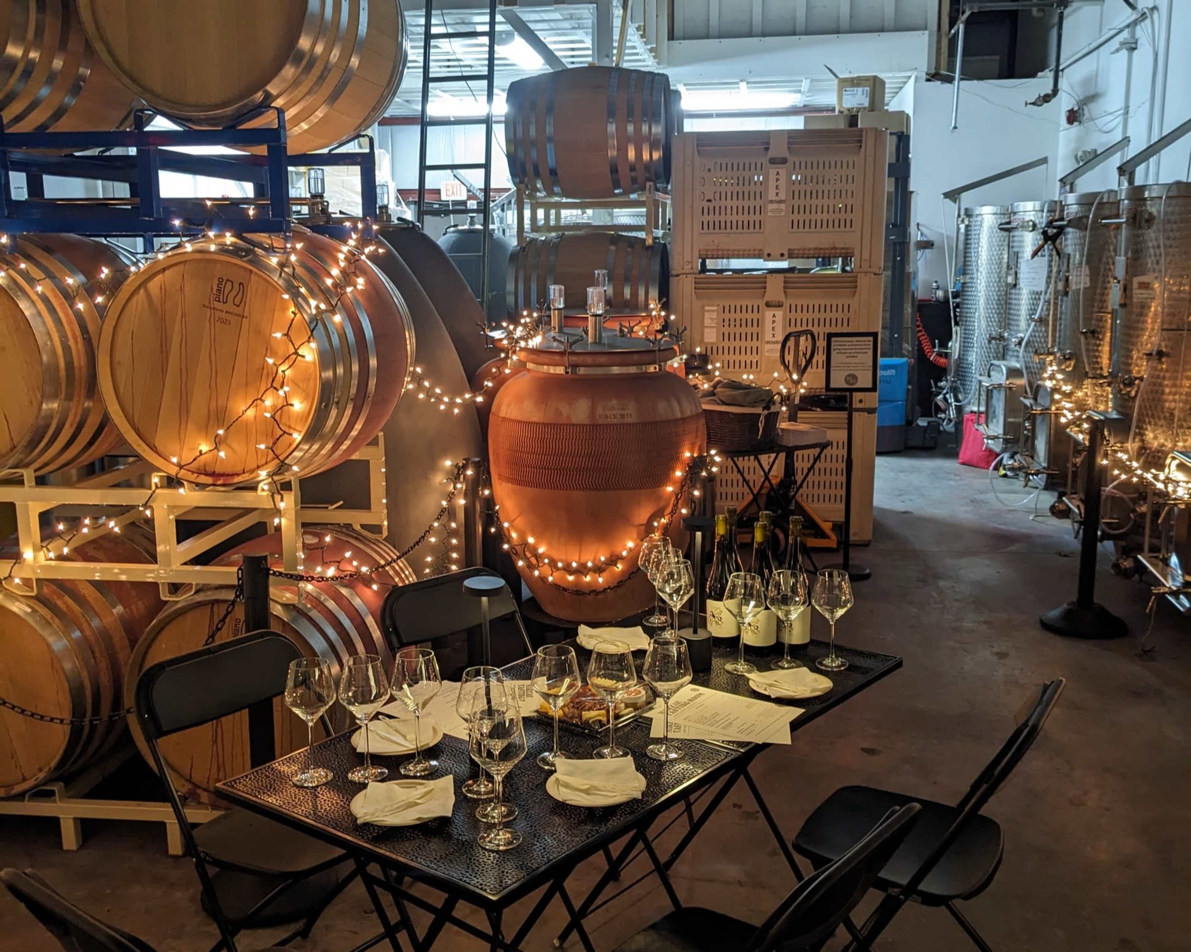 A table set for a wine tasting is positioned among barrels and fermentation tanks in a winery.