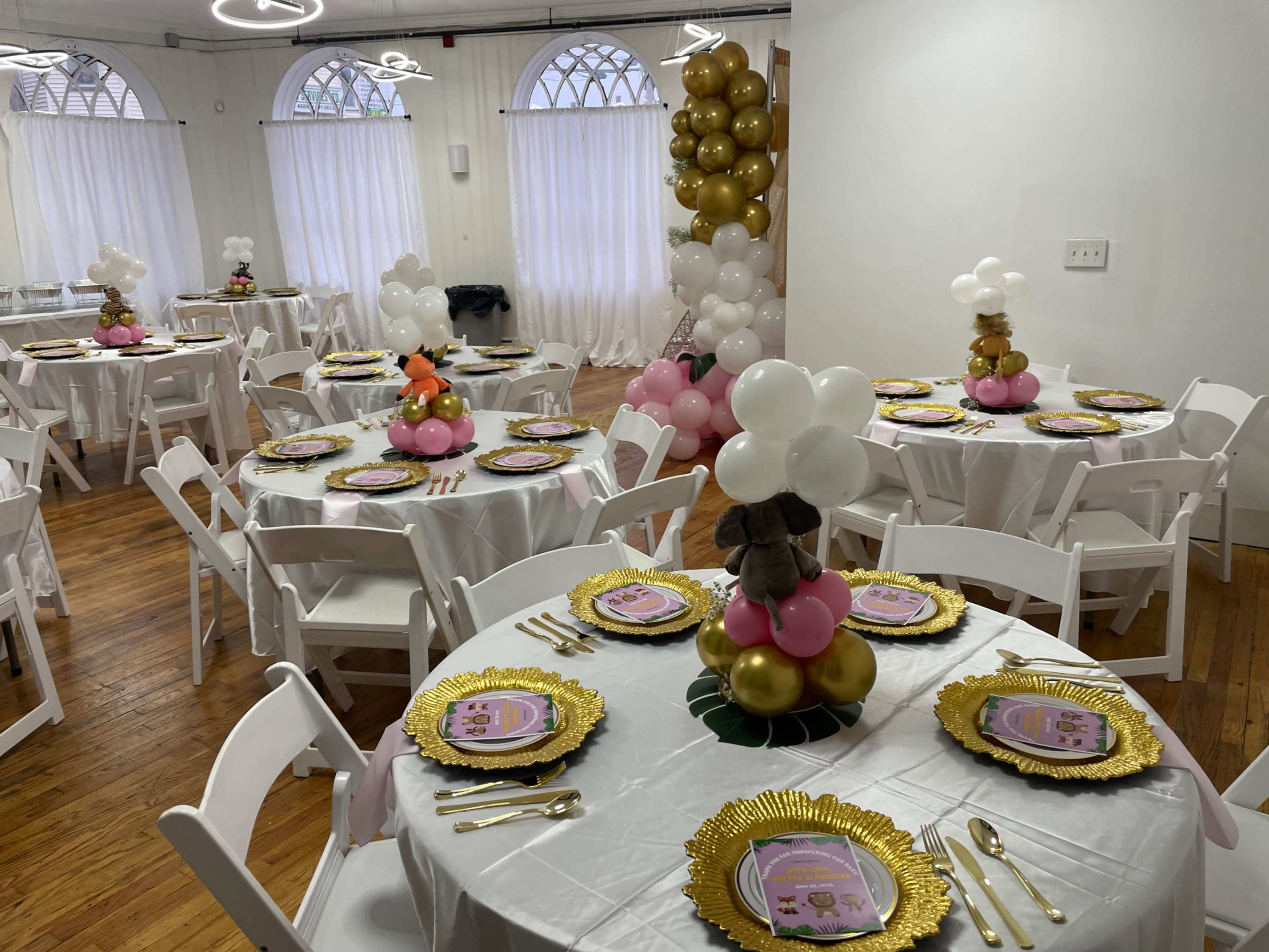 A banquet hall with several round tables set with gold-rimmed plates, decorative balloons, and centerpieces featuring stuffed animals and colorful balloons.