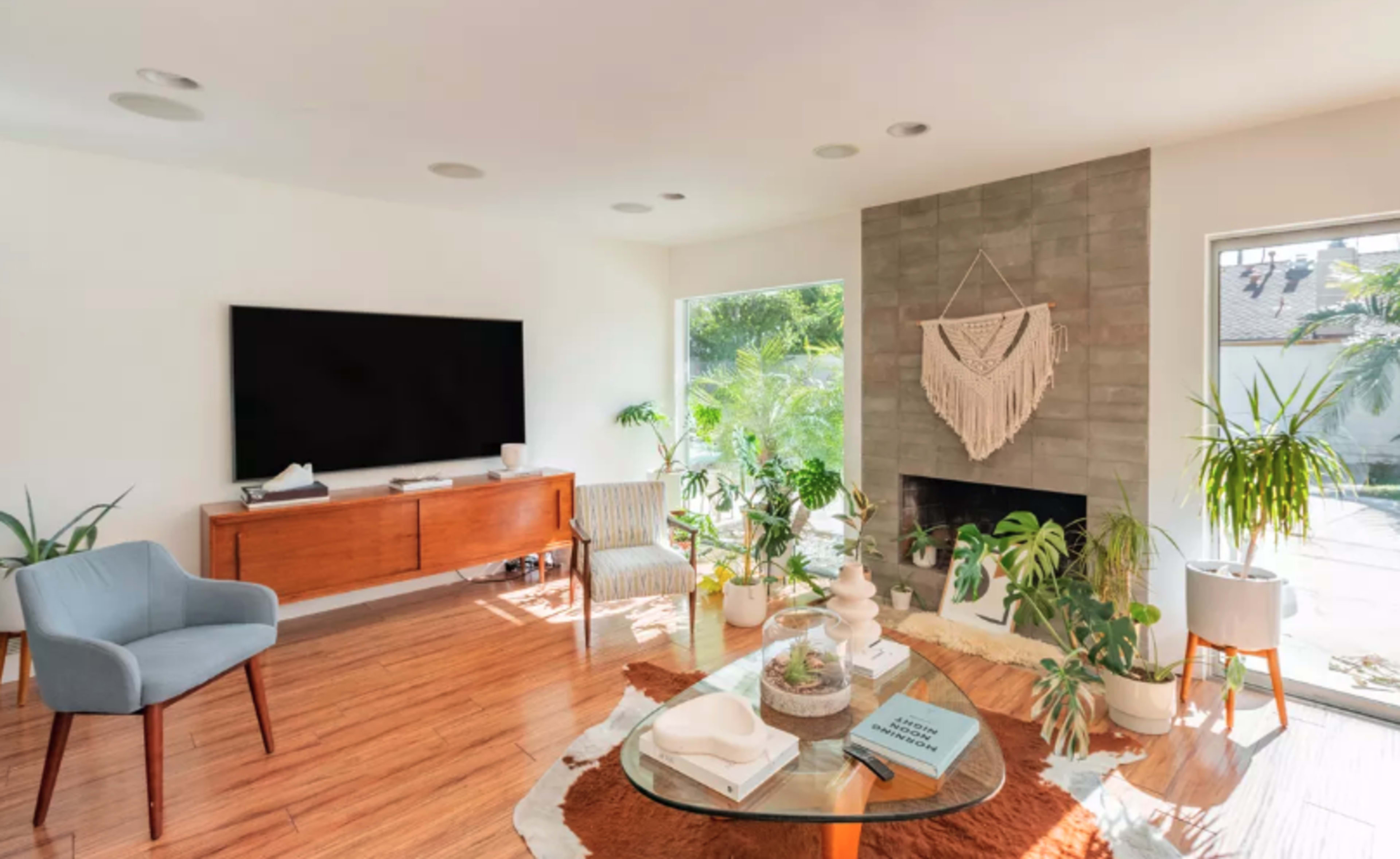 A modern living room features a wall-mounted television, a wooden media console, and large windows surrounded by various indoor plants.