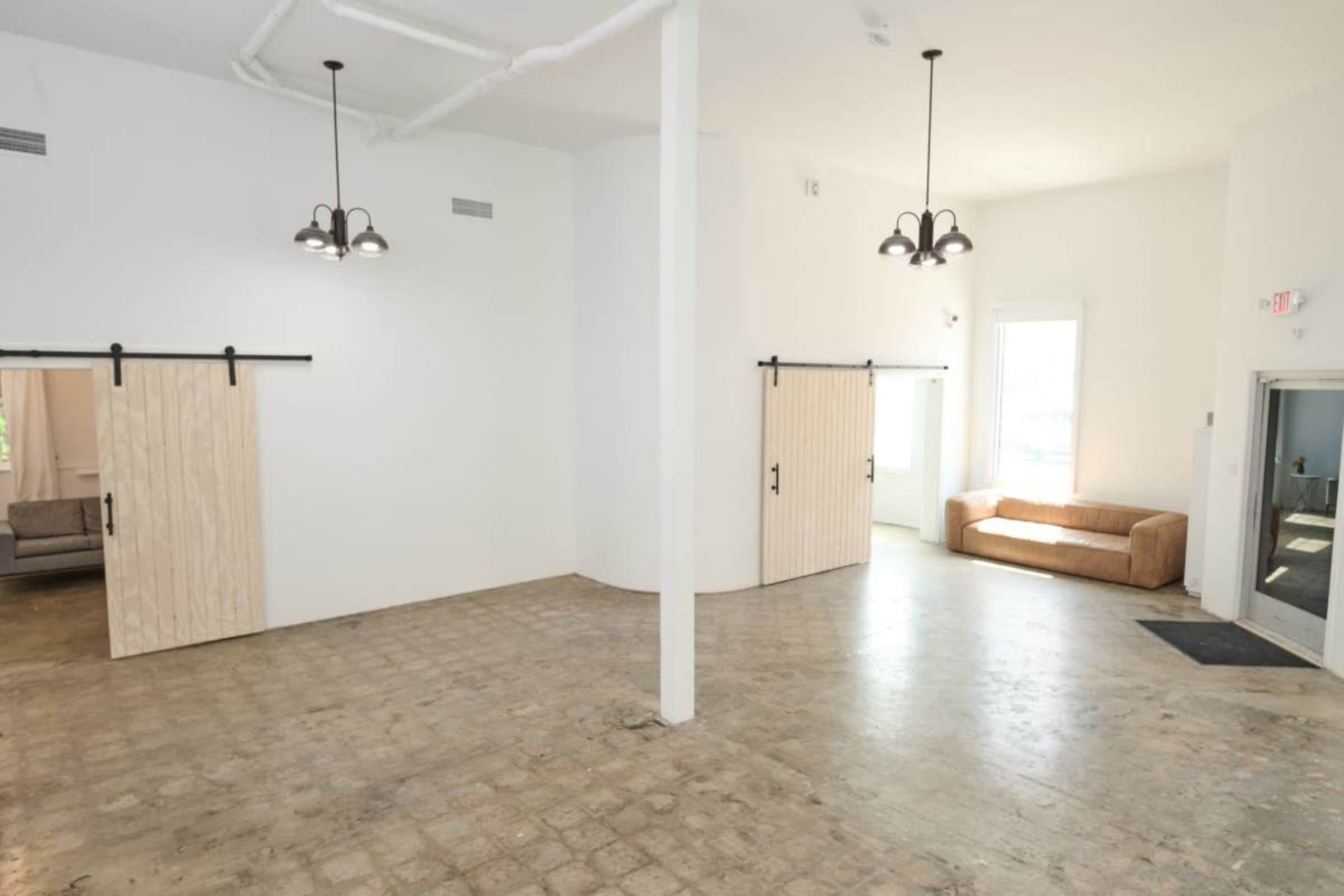 The image shows a spacious, empty room with white walls, concrete flooring, and sliding barn doors on either side, along with a couch positioned near a window.