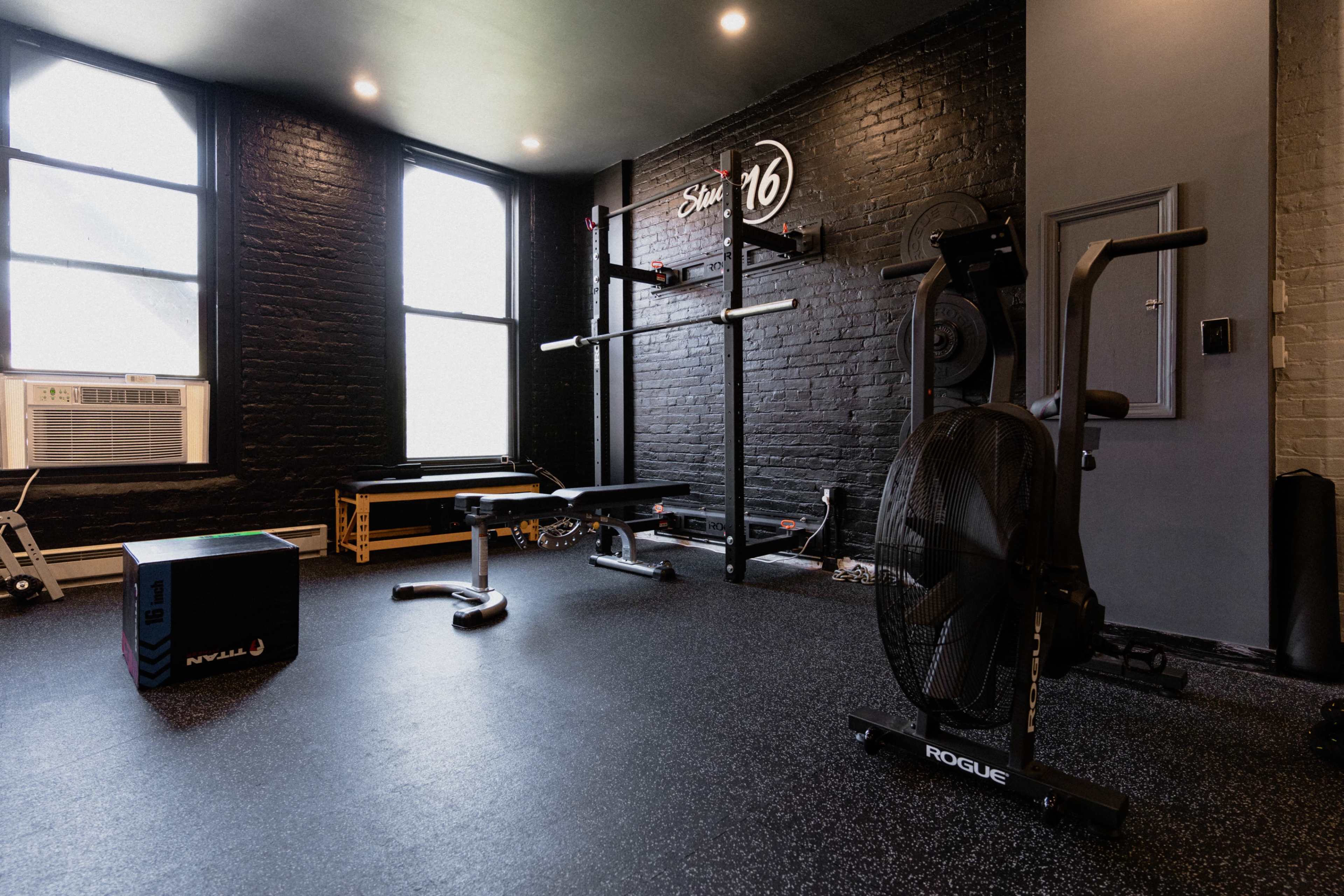 A modern gym interior with black walls, large windows, an air conditioning unit, and various exercise equipment including a rowing machine and a weight bench.