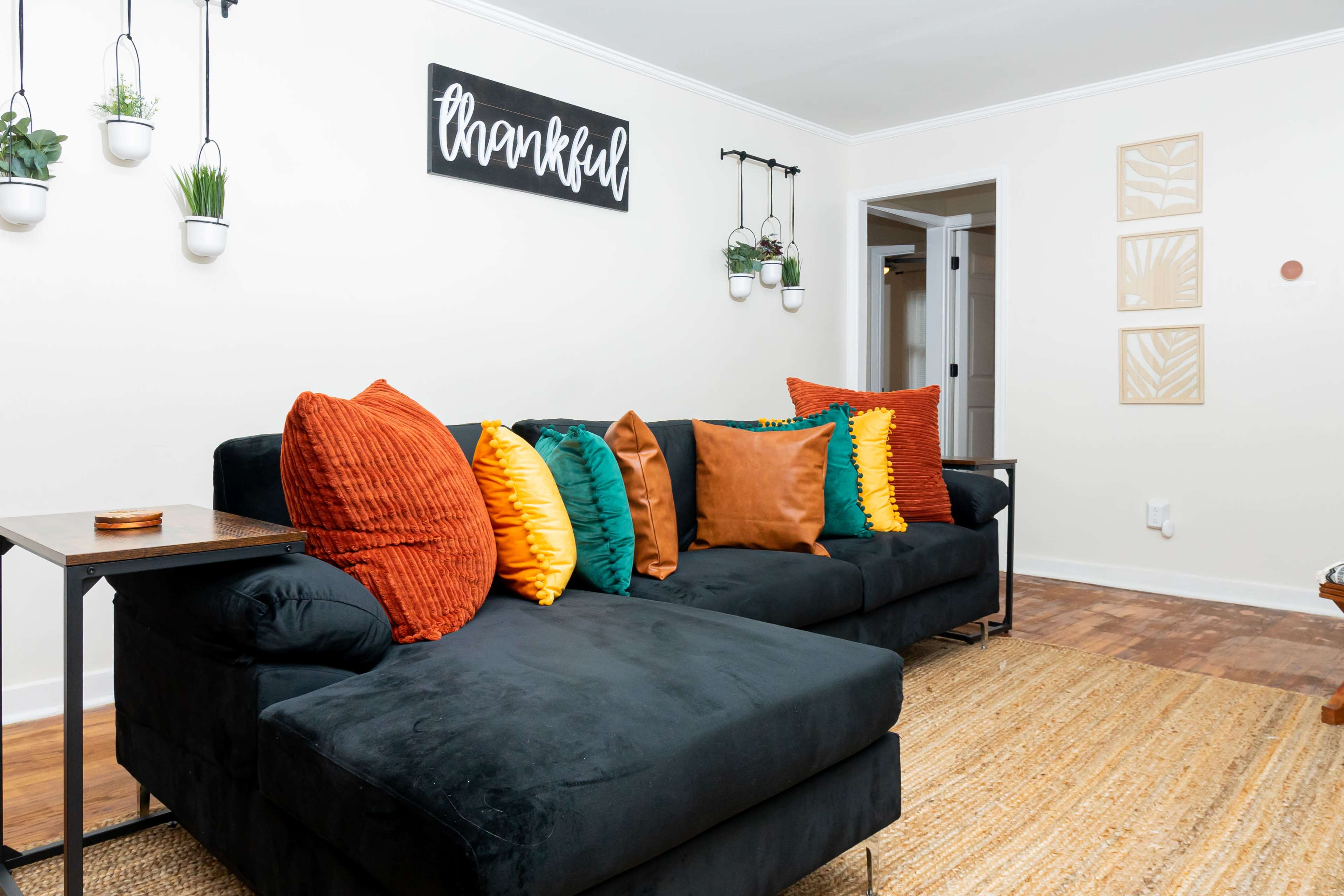 A black sectional sofa with various decorative pillows is positioned against a light-colored wall adorned with planters and artwork.