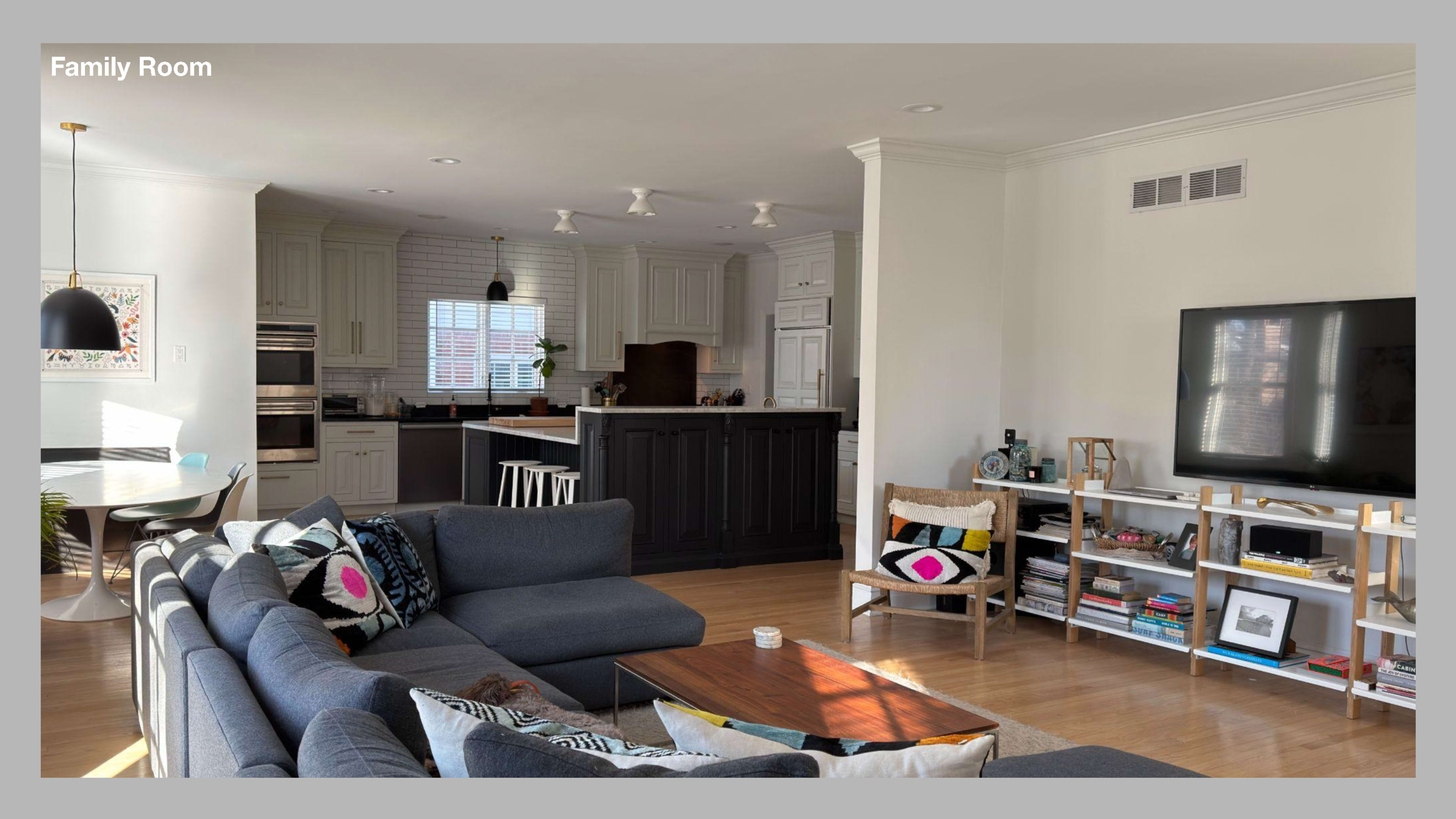 The image shows a modern family room featuring a gray sectional sofa, a wooden coffee table, and a television on a white cabinet, with an open kitchen visible in the background.