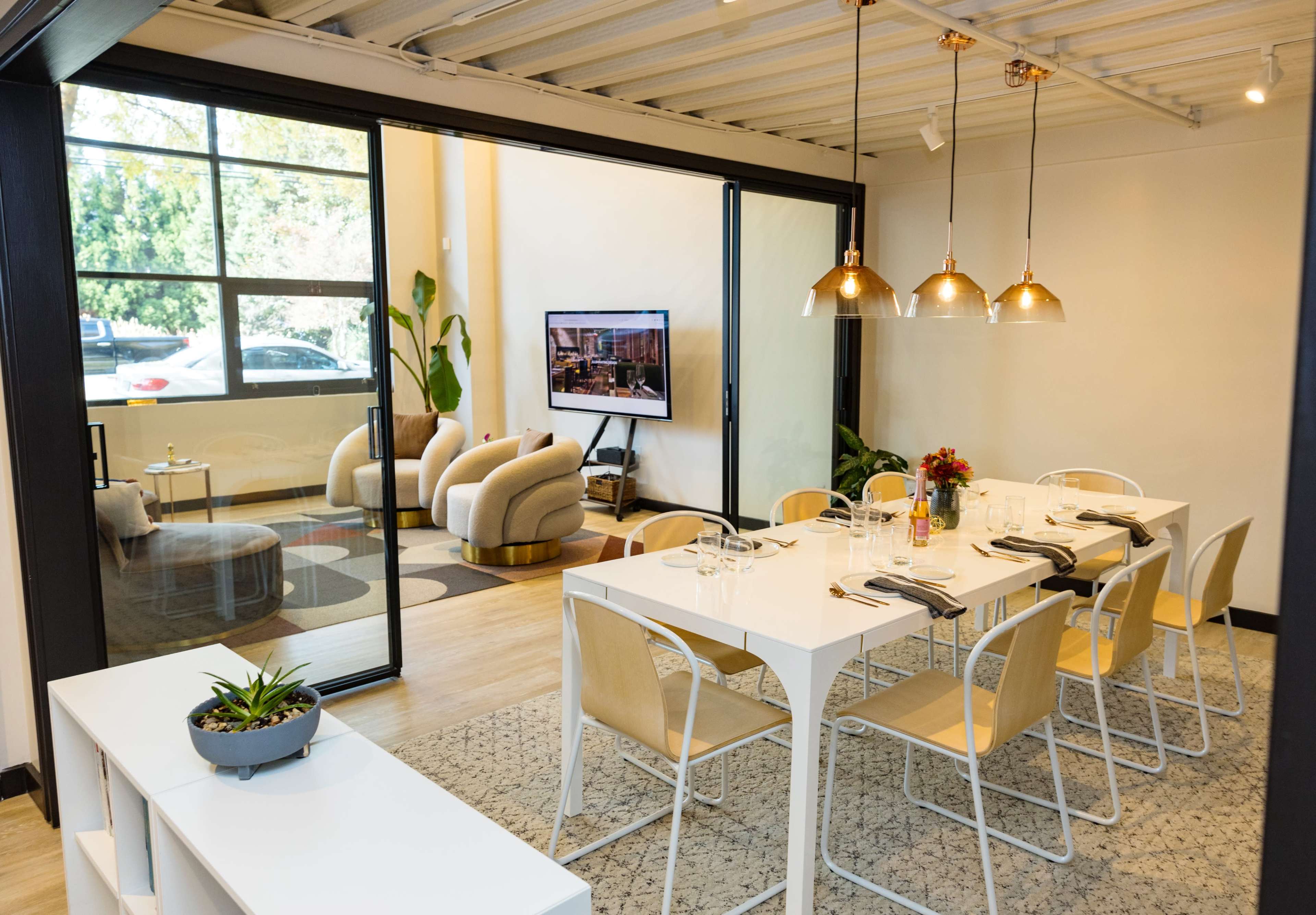 A modern dining area with a long white table set for a meal, alongside a cozy seating area visible through a glass partition.