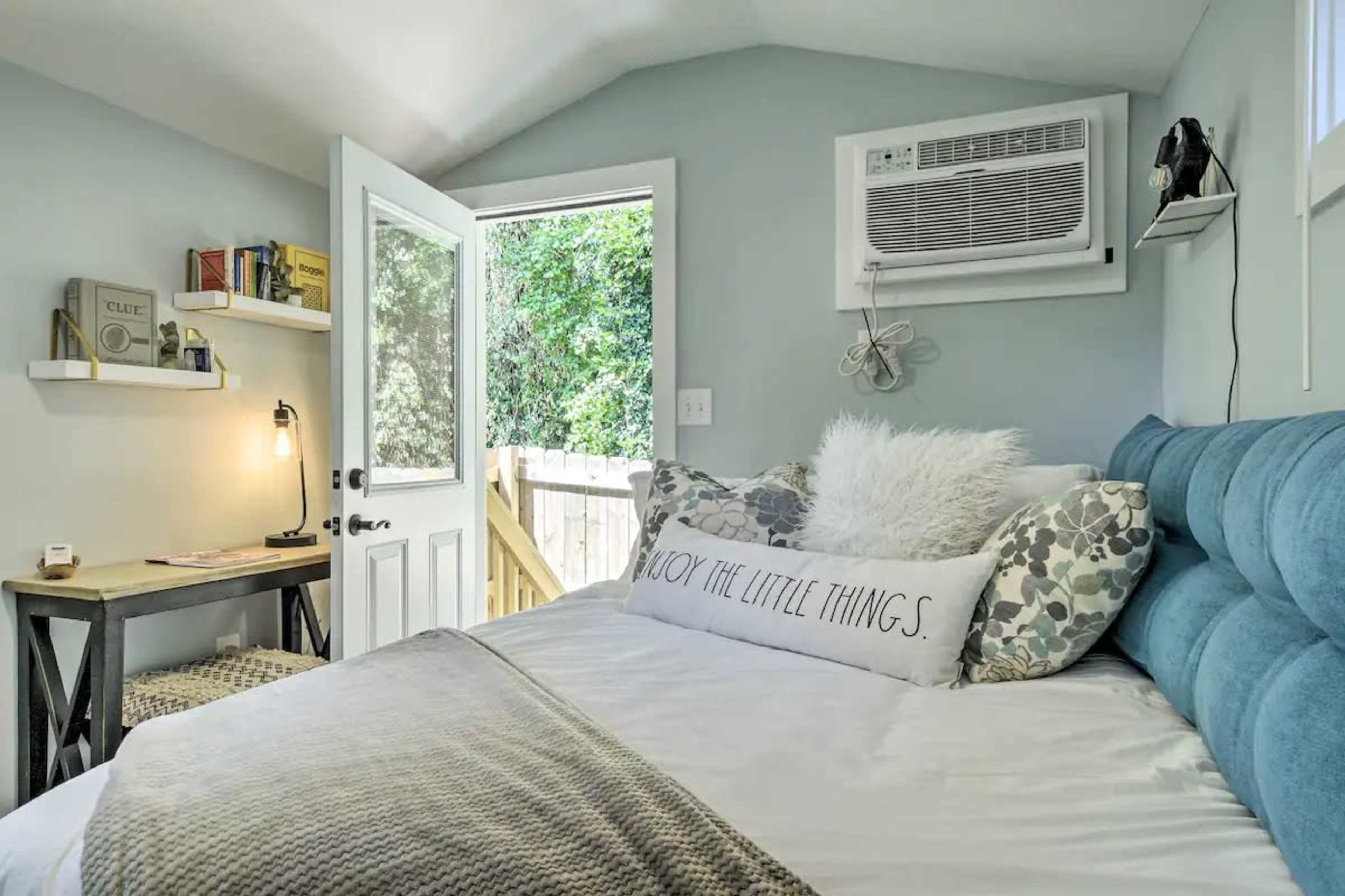 A small bedroom with a blue upholstered headboard, a bed with decorative pillows, a study table, and an air conditioning unit, along with a door leading to an outdoor area.