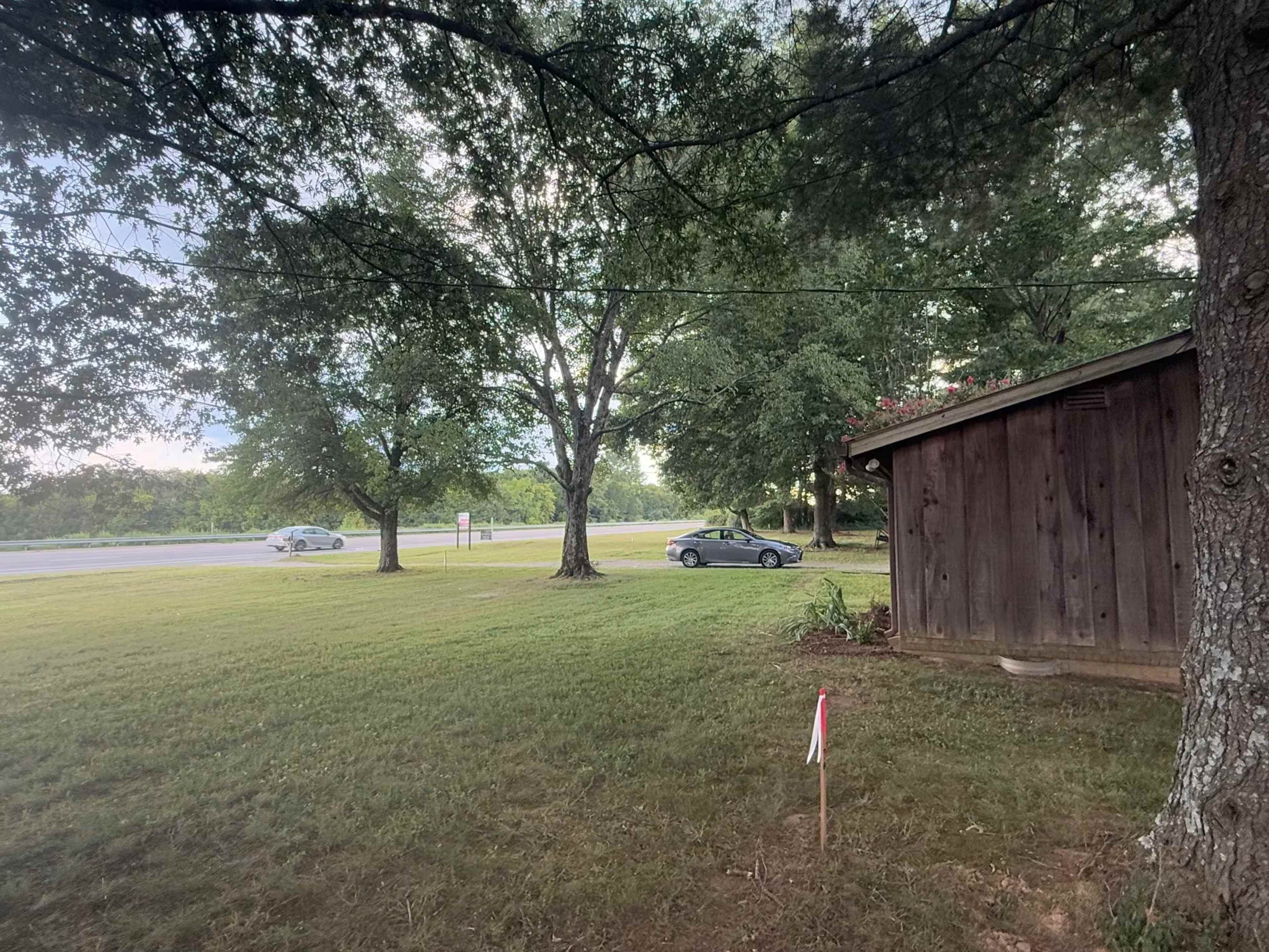 A view of a grassy area beside a road, featuring a wooden structure and a parked car in the distance, with trees providing shade.