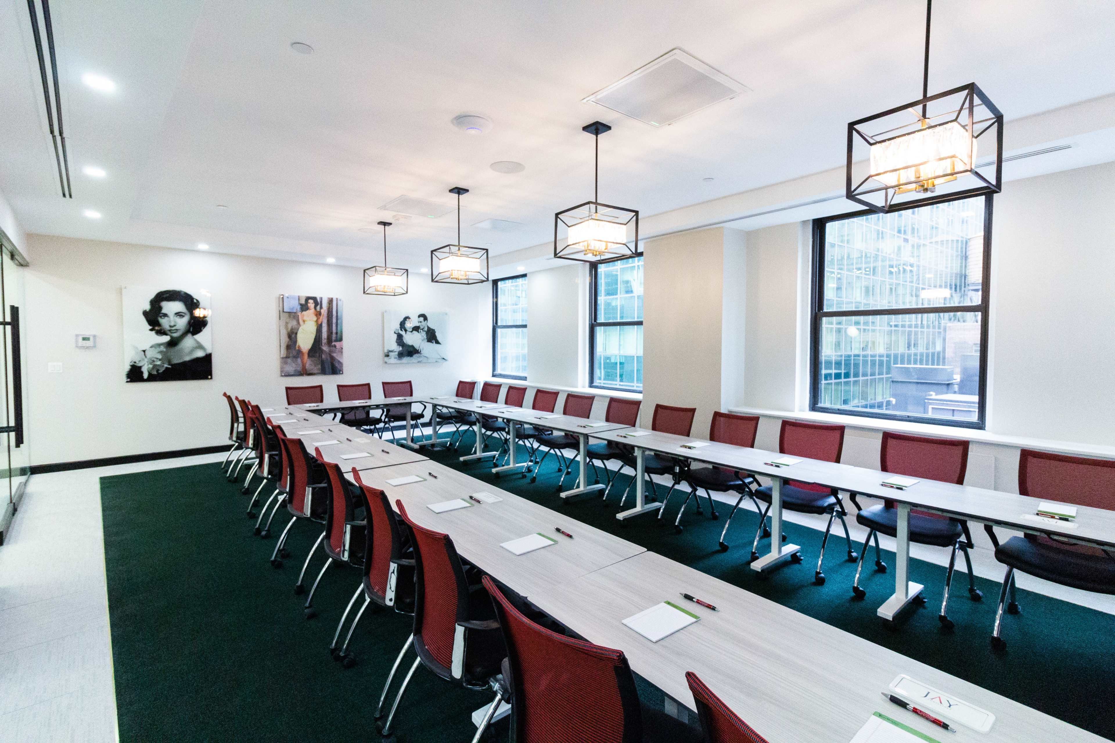A modern conference room features long tables arranged in a U-shape, equipped with chairs and stationery, illuminated by overhead pendant lights.