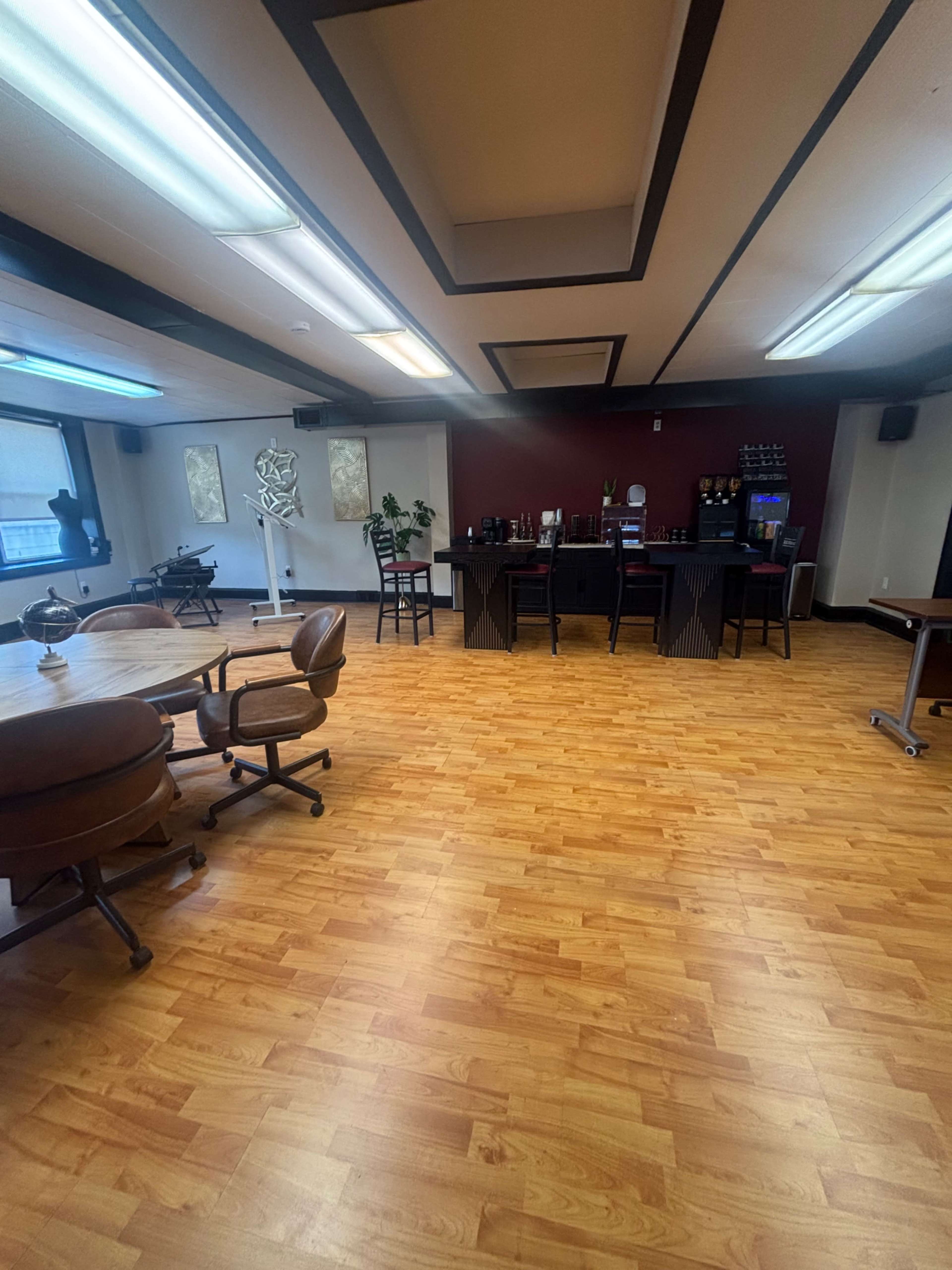 The image shows a spacious, modern office interior with wooden flooring, a bar area against a red wall, and several seating arrangements.