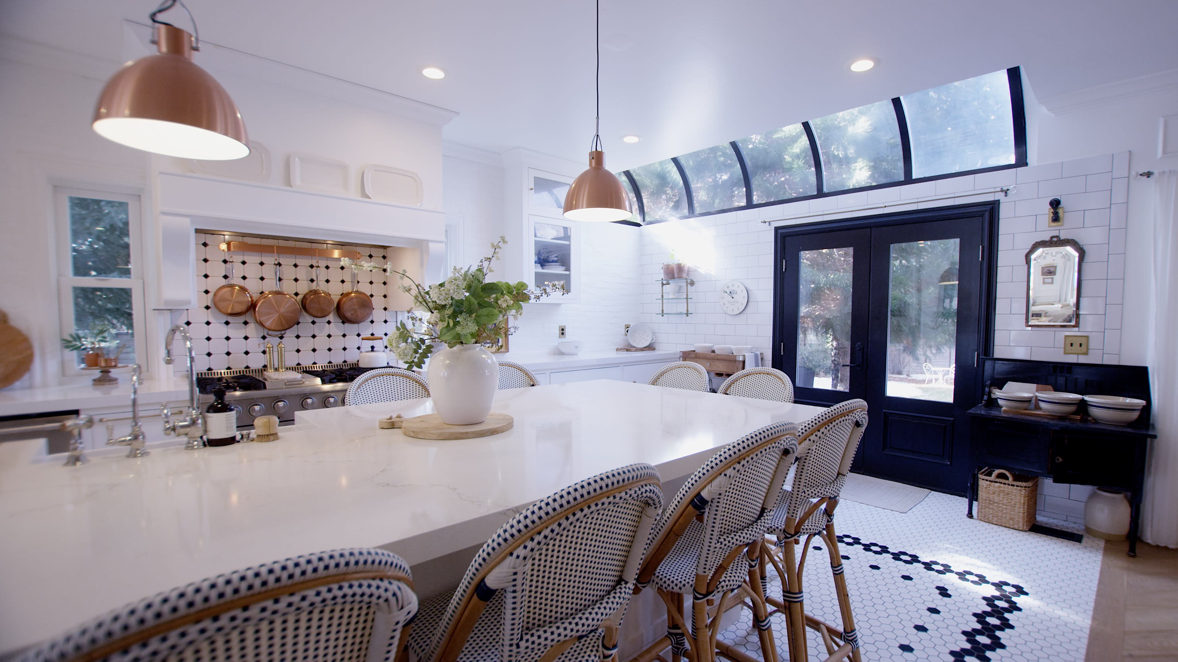 A spacious kitchen features an island with high-backed chairs, wooden cookware displayed on the wall, and a large window allowing natural light to fill the space.