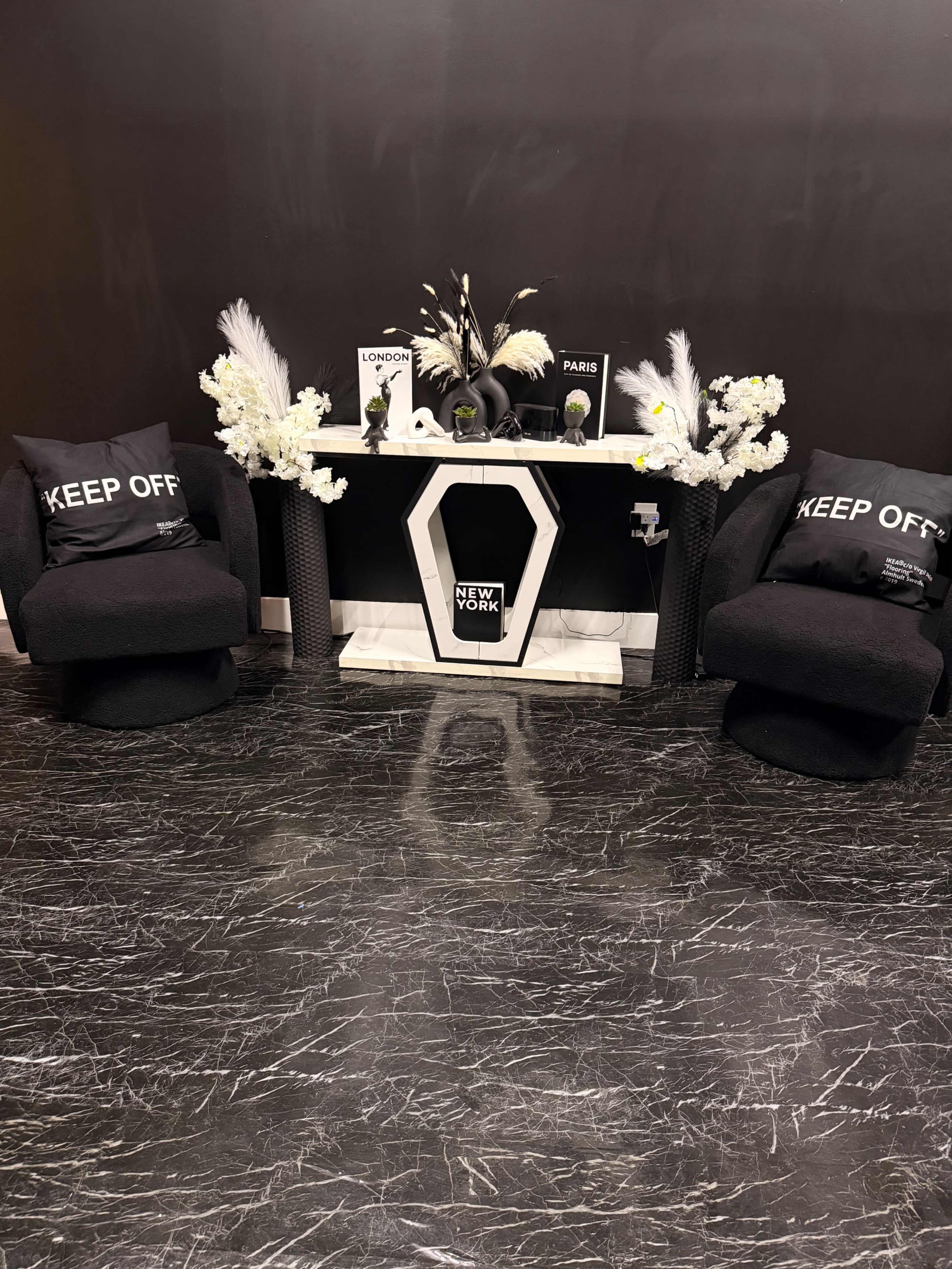 The image shows a modern seating area with two black chairs, a decorative table featuring plants and city-themed decor items, all set against a dark wall and floor.