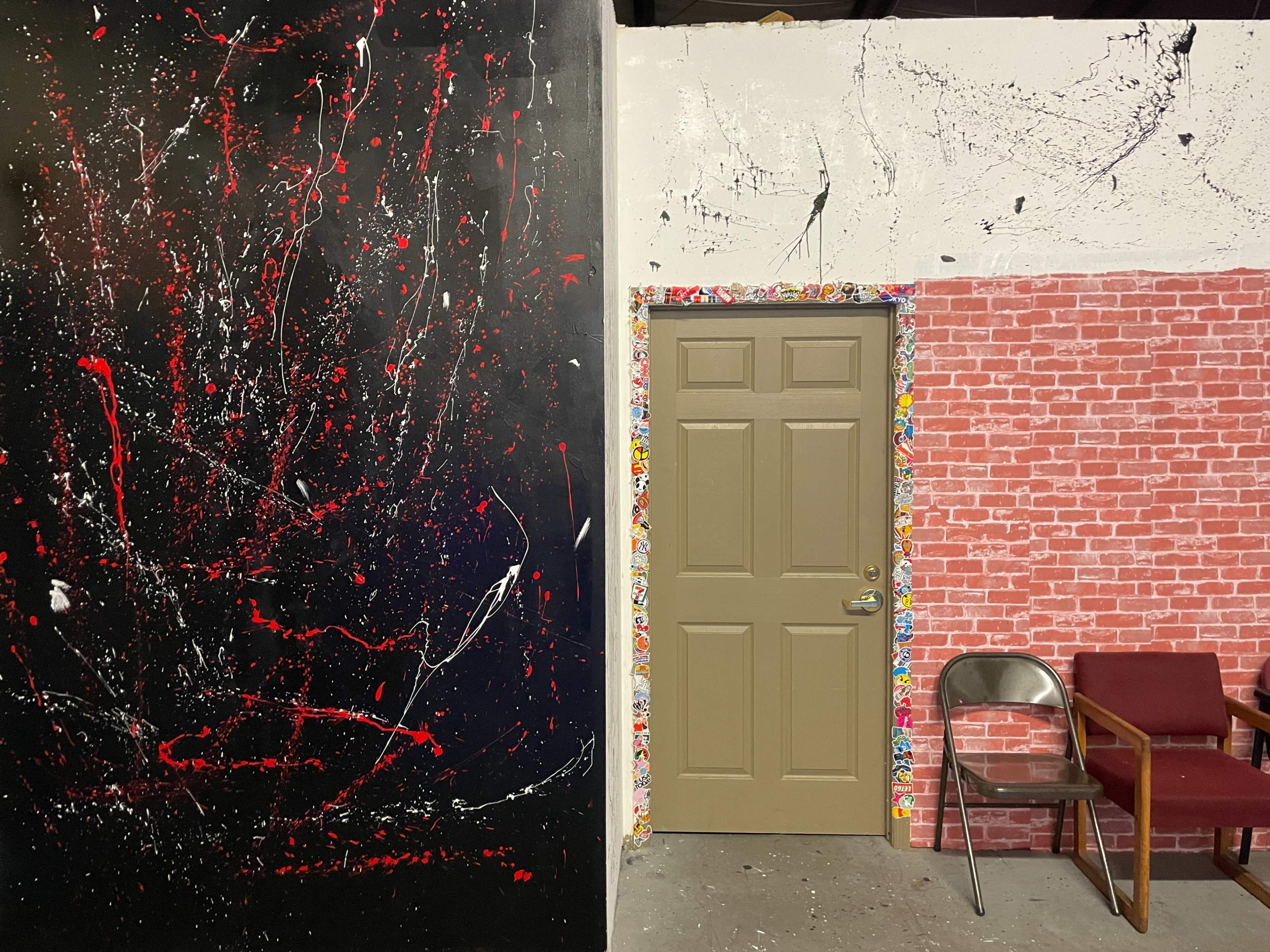 A room with a black wall featuring abstract paint splatters, a beige door framed by colorful wallpaper, and a red-brick wall adjacent to a metal chair and a maroon armchair.