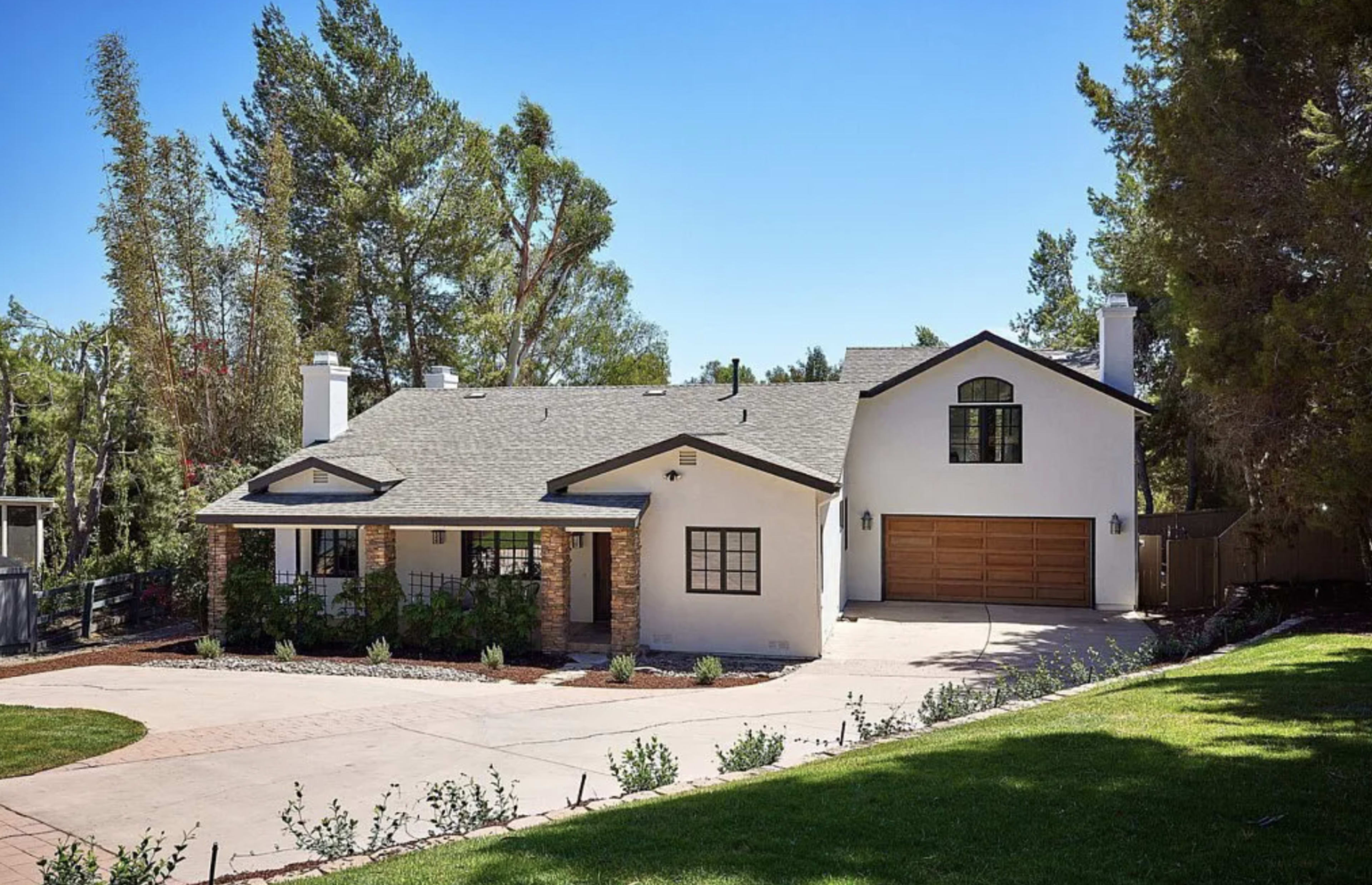A large, two-story house with a gray roof and white exterior is situated on a landscaped lot with a circular driveway and garage.
