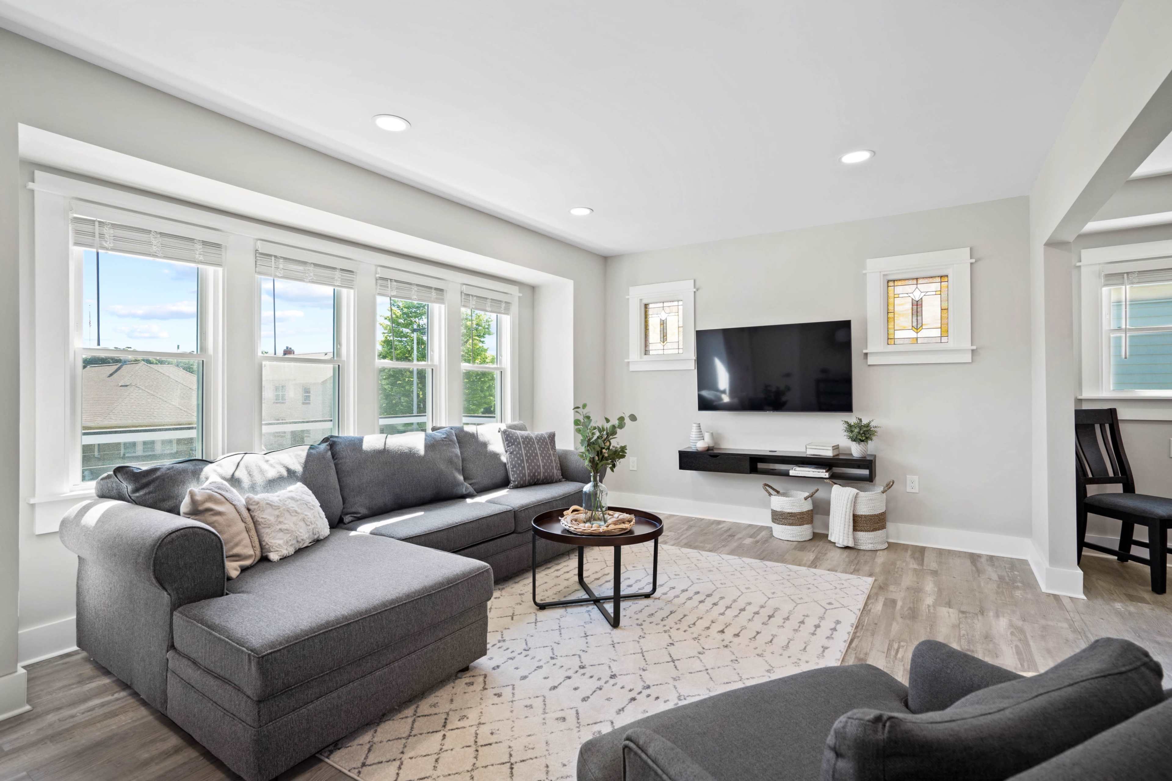 A modern living room with a sectional sofa, a coffee table, and a mounted television on a light-colored wall, illuminated by natural light from large windows.